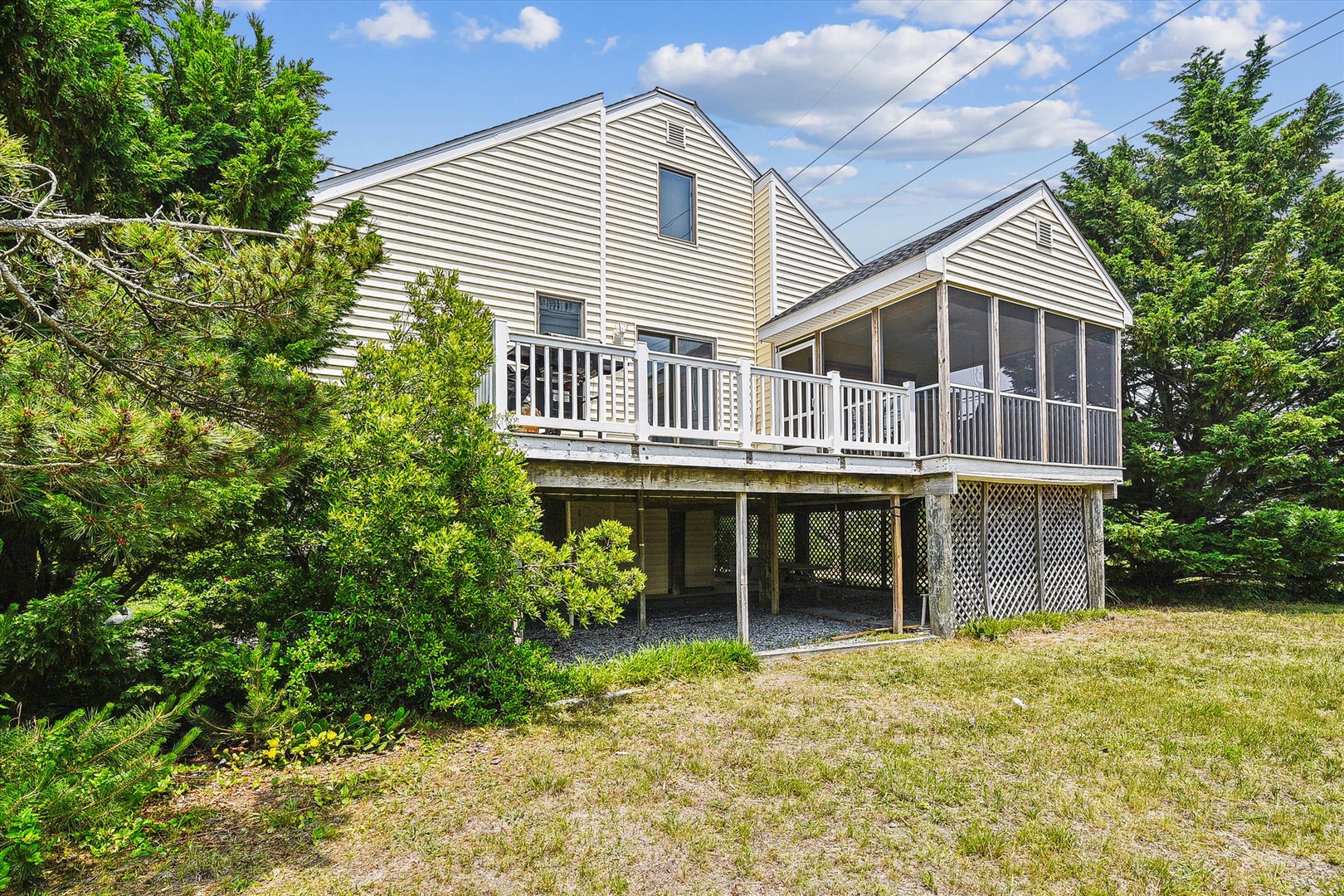 Classic beach house with elevated deck, screened porch, and covered parking, nestled among coastal greenery under summer skies. A peaceful retreat near the shore.