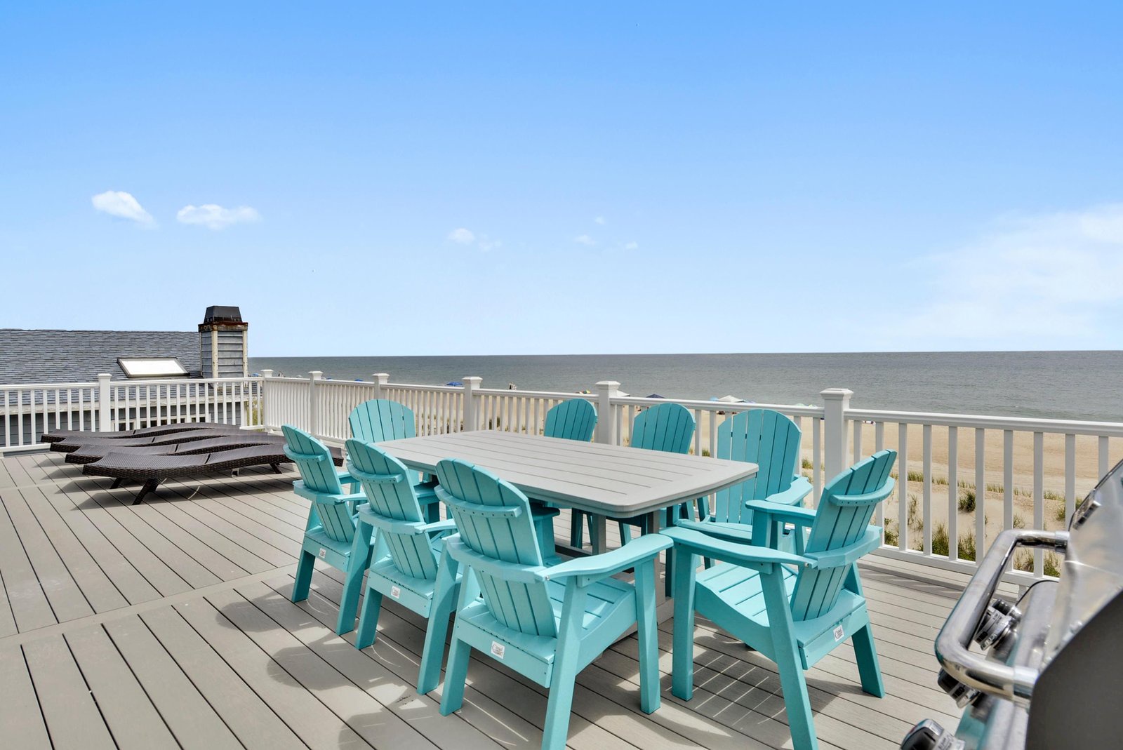 A spacious rooftop deck featuring turquoise Adirondack chairs around a dining table, perfect for oceanfront meals and relaxation.