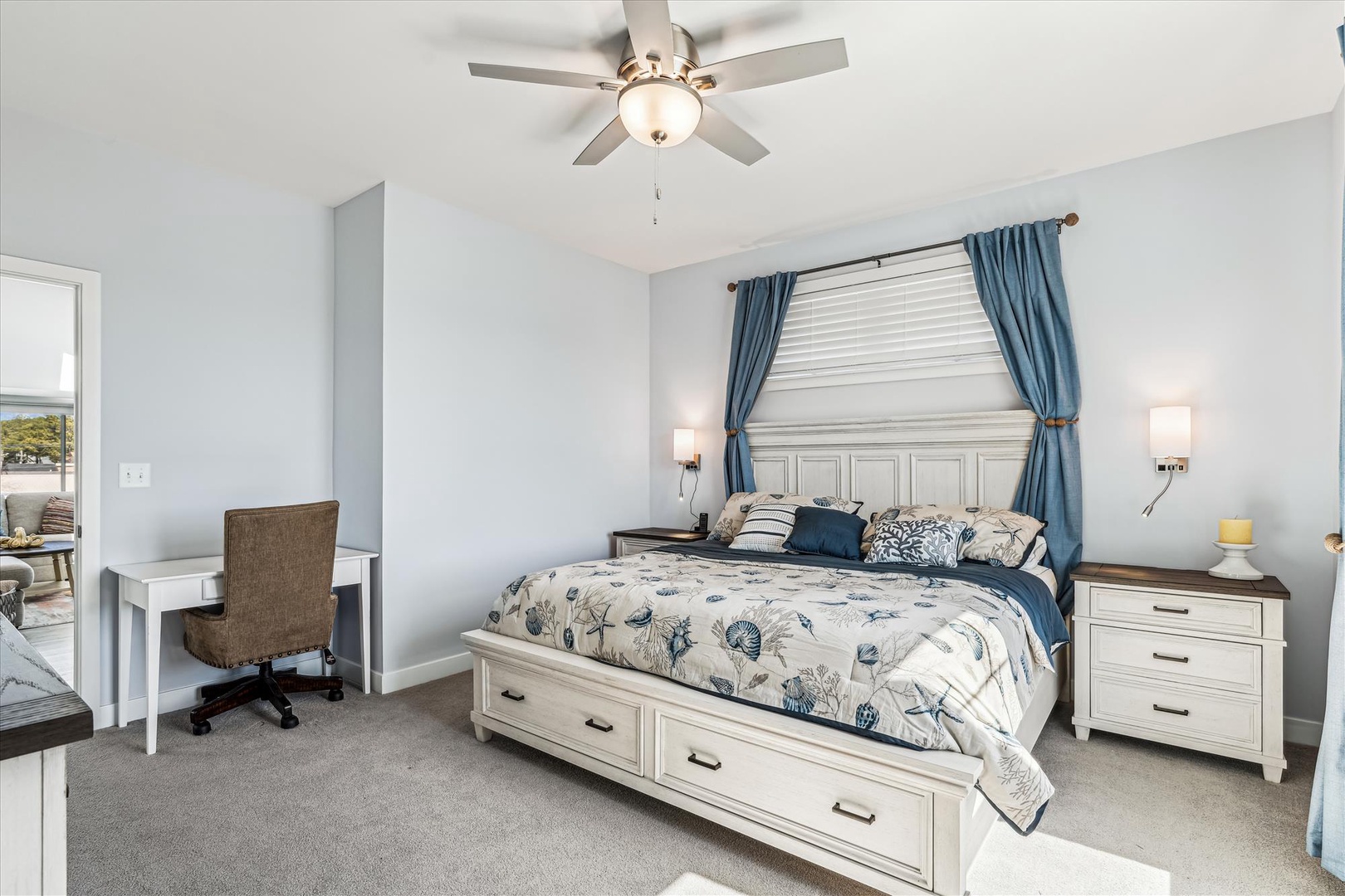 Unwind in your serene bedroom featuring a plush queen bed with coastal-inspired linens, soft blue curtains, and a gentle ceiling fan to keep you cool.