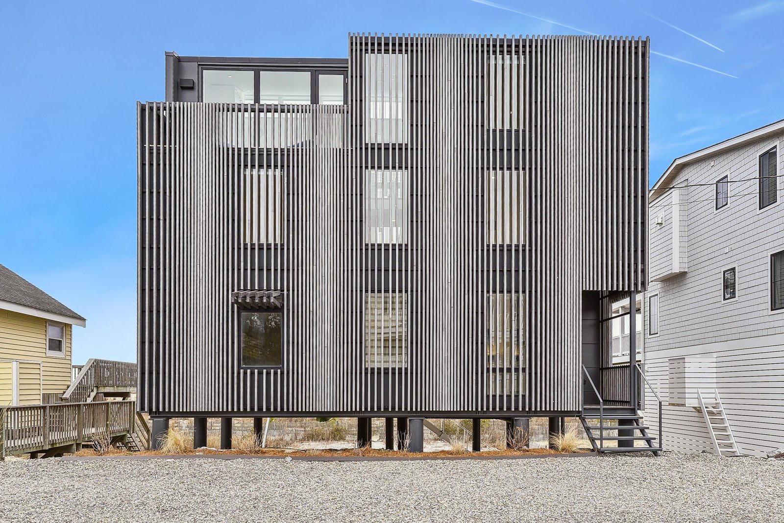 Modern architectural design featuring distinctive vertical siding, set in a coastal neighborhood with neighboring beach homes and ample gravel parking.