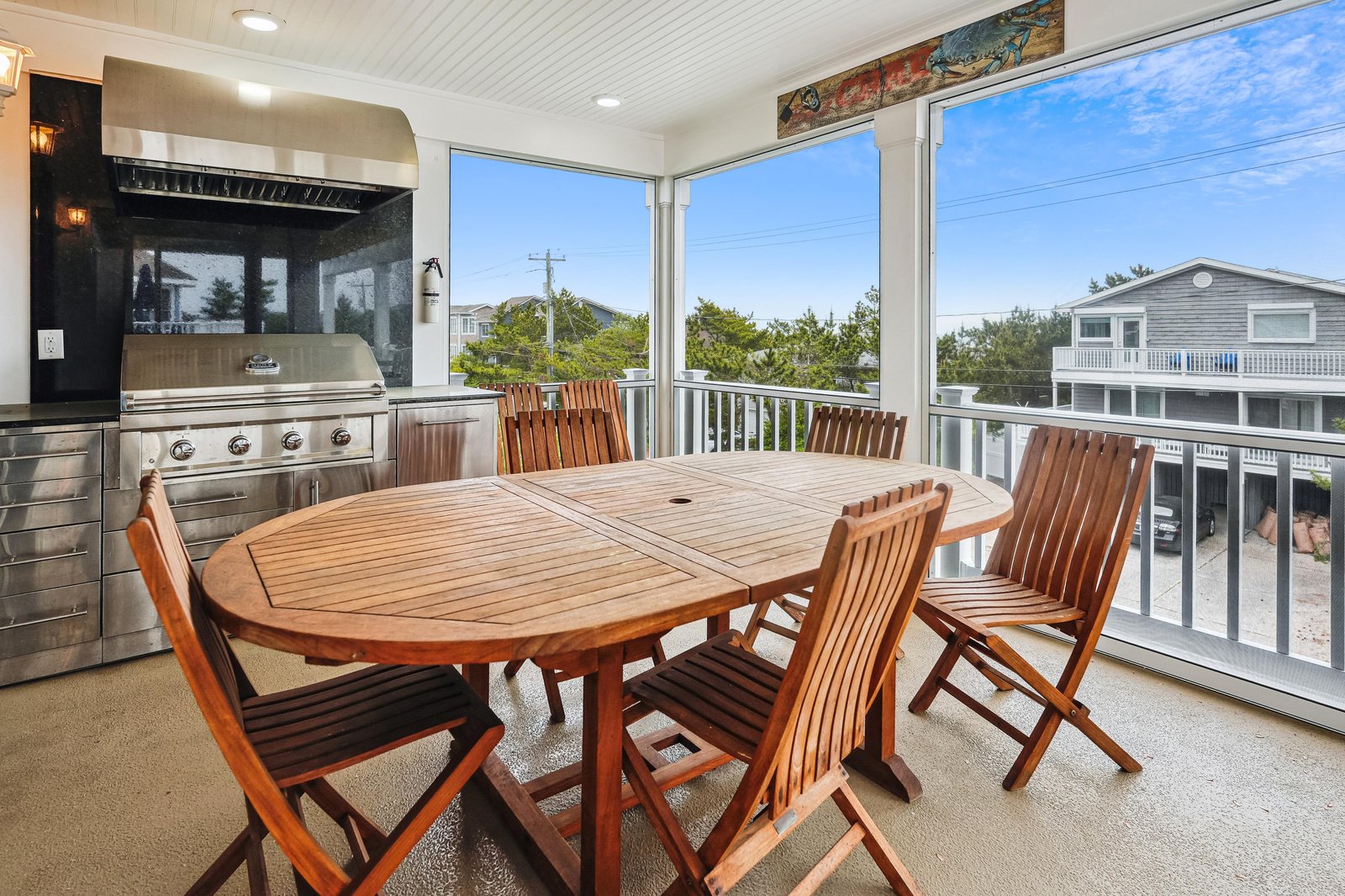 Private screened porch with floor-to-ceiling windows, wooden dining table for eight, built-in grill, and views of lush greenery and nearby beach homes.
