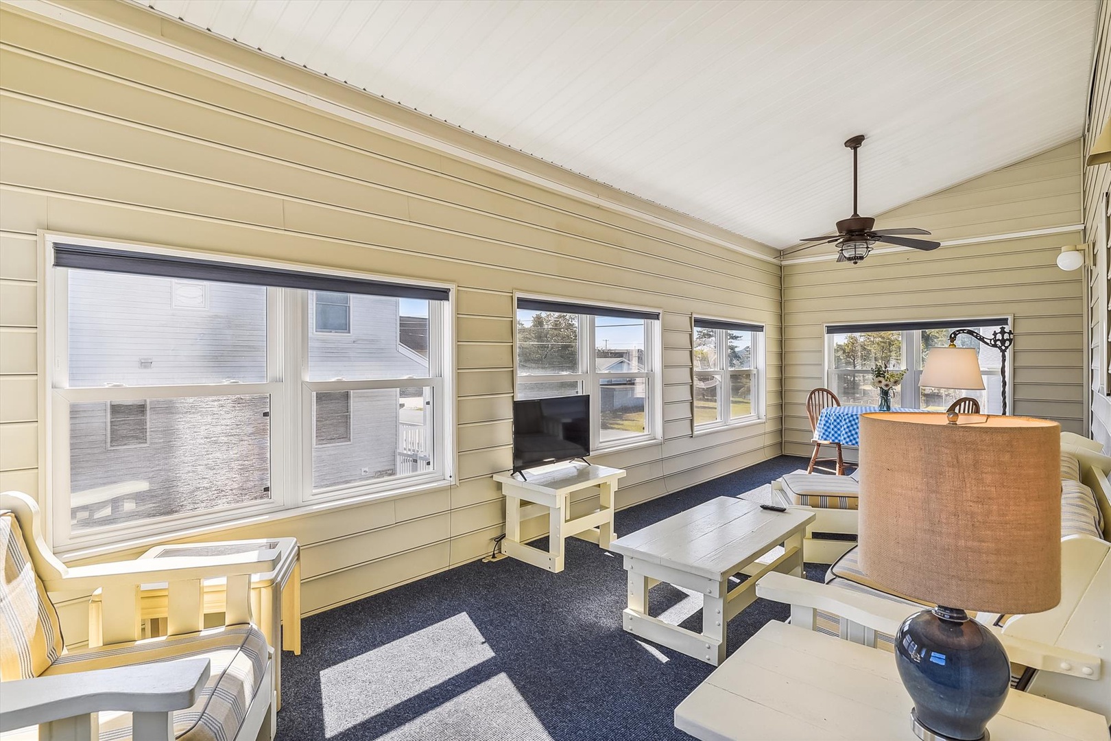 Your sun-drenched living room features coastal charm with whitewashed furniture, a ceiling fan, and panoramic windows framing serene water views—perfect for relaxing after beachside adventures.