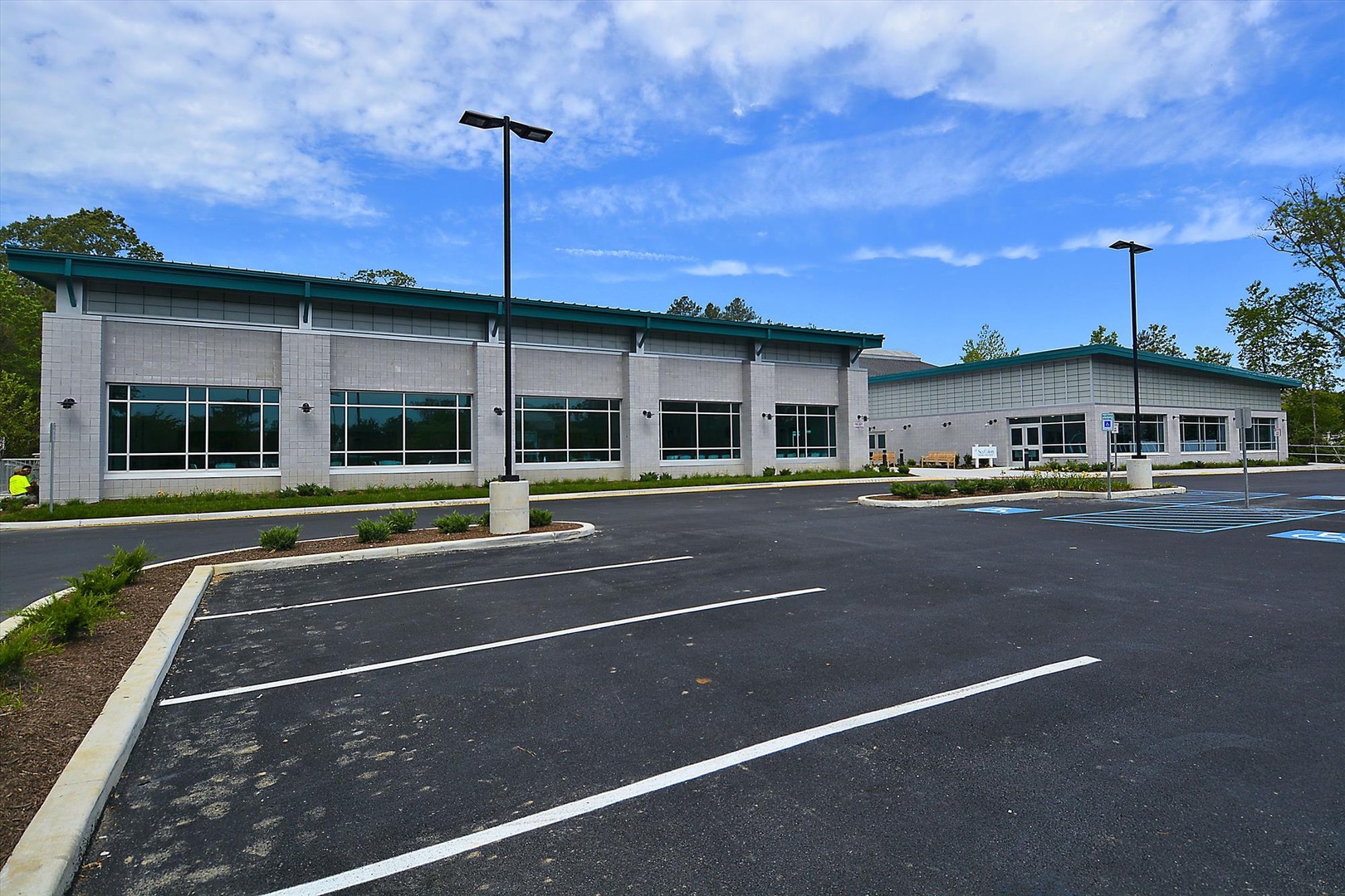 Modern commercial building featuring a distinctive green metal roof and large windows, set against landscaped grounds with ample parking and surrounded by natural wooded areas.