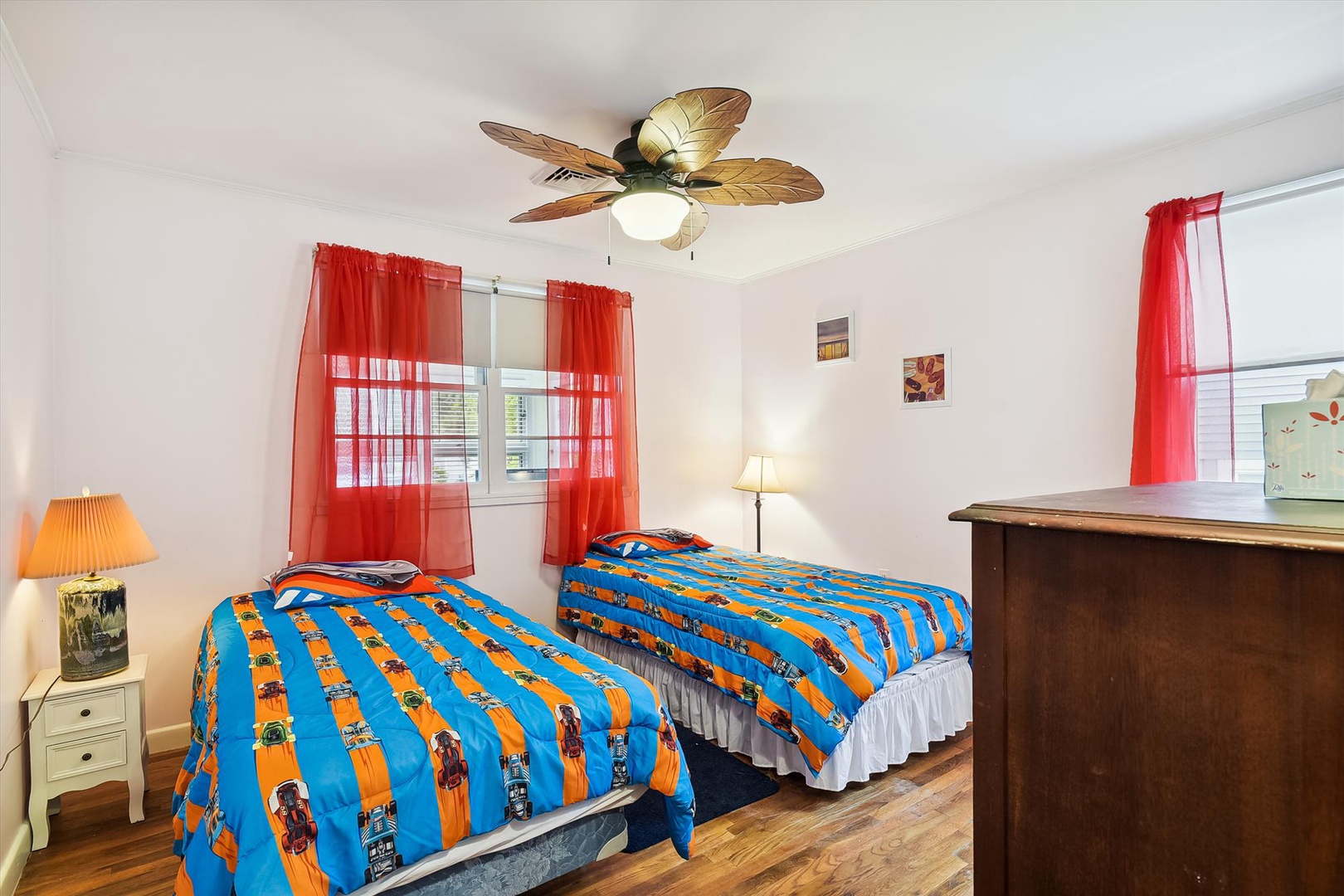 Unwind in this cheerful bedroom with twin beds dressed in vibrant linens, a tropical ceiling fan, and breezy red curtains that frame natural light—your peaceful retreat awaits.