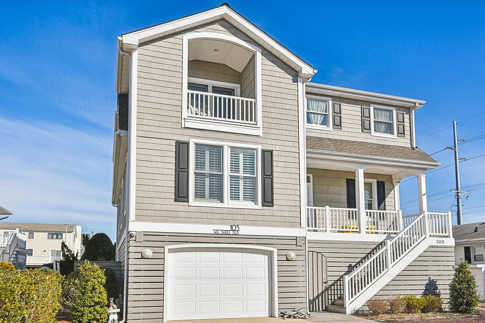 A modern coastal home features classic gray shingle siding, white railings, and multiple balconies under clear blue skies in a quiet residential neighborhood.