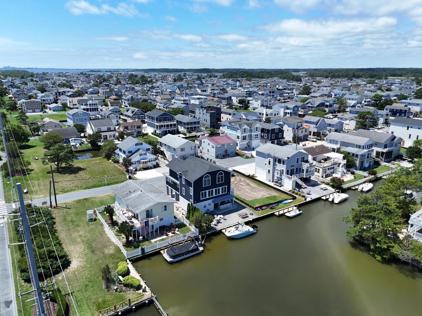 Aerial view of a charming waterfront neighborhood, with the property located among coastal homes and boat docks along a calm canal, surrounded by lush greenery and open skies.