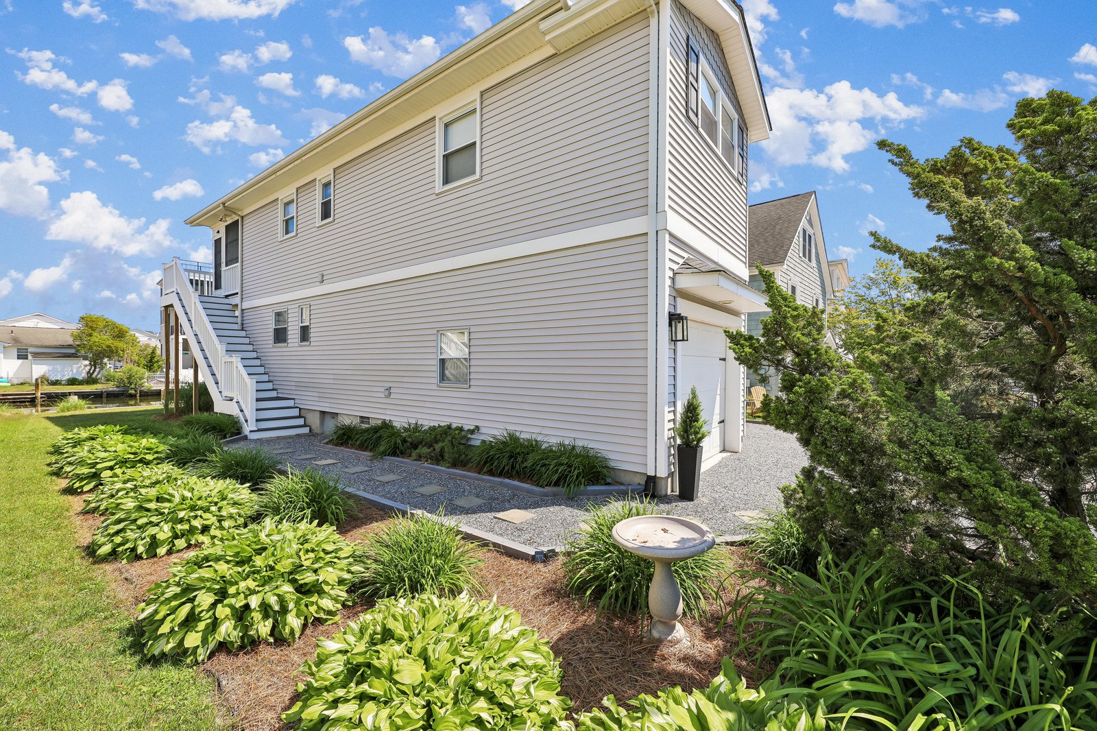 A charming two-story coastal retreat featuring a landscaped garden with lush hostas and a peaceful birdbath, offering a welcoming arrival experience beneath sunny skies.