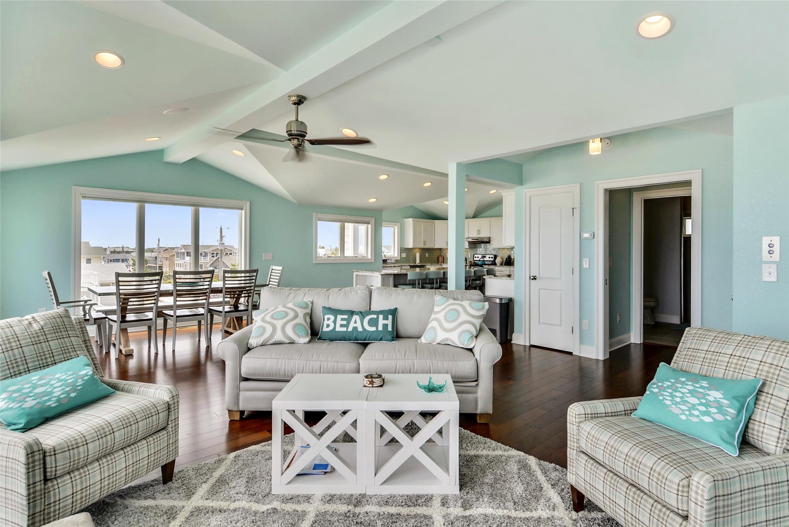 Relax in your bright, airy living space with cheerful coastal colors, comfortable seating, and a ceiling fan overhead. Sunlight pours in through large windows, creating a welcoming atmosphere for unwinding after beach adventures.