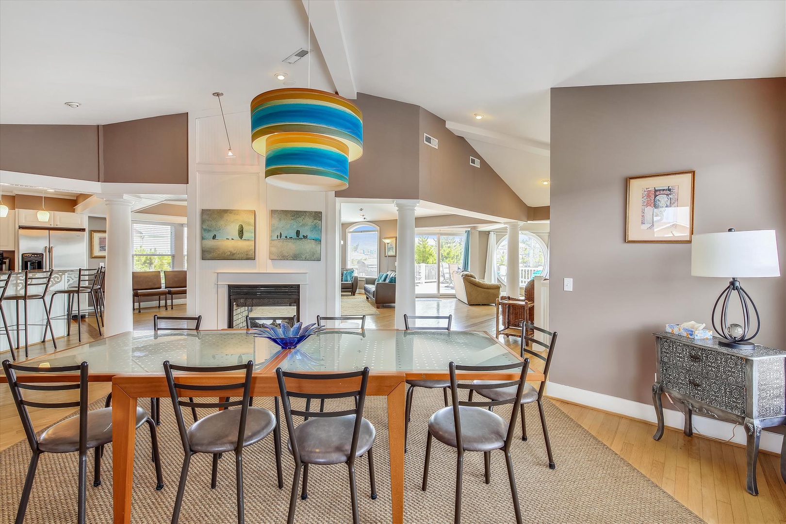This bright, open living and dining space features soaring vaulted ceilings, abundant natural light, and a striking blue-striped chandelier that adds playful coastal charm to your vacation home.