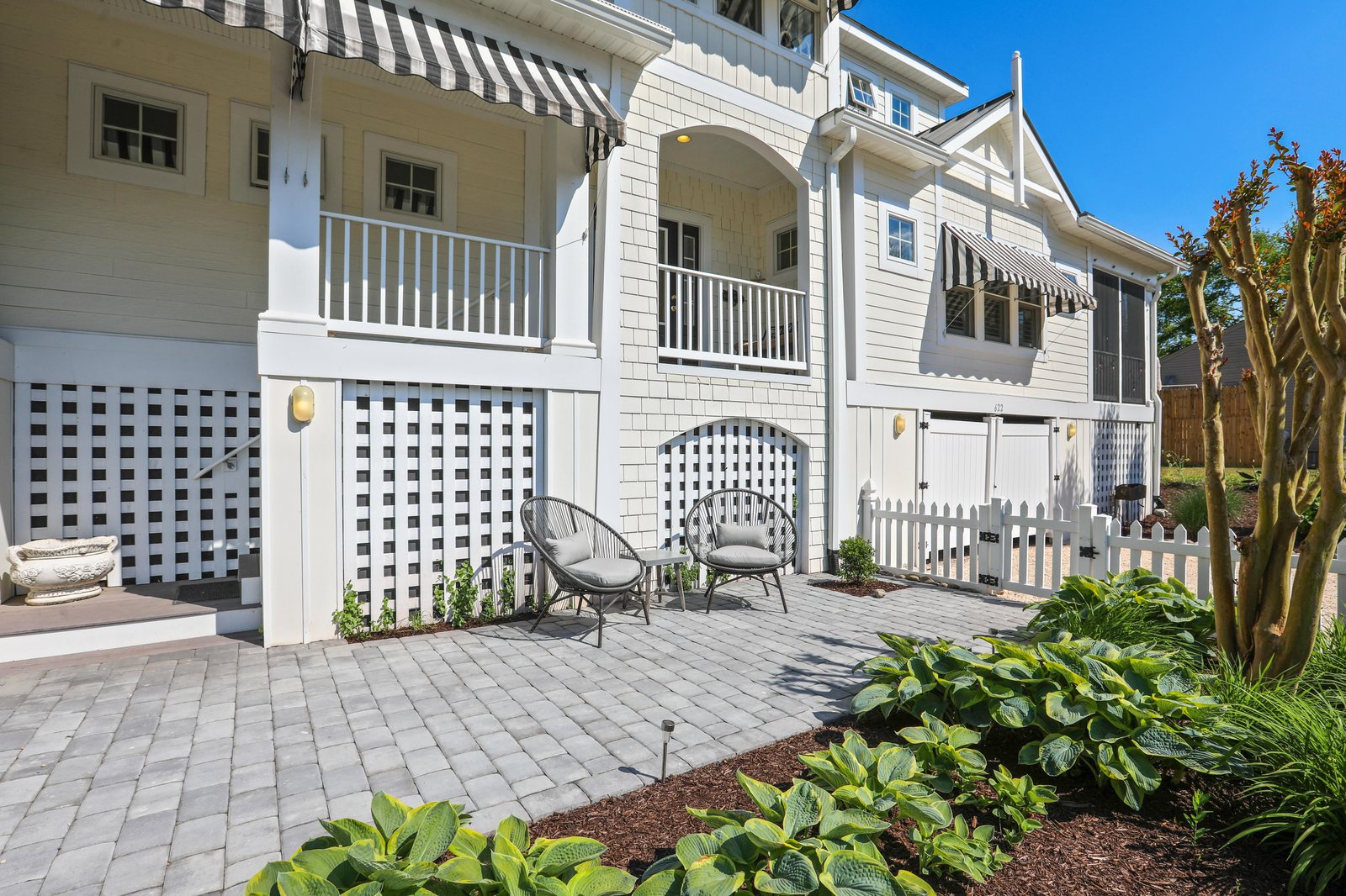 This bright and cheerful property greets you with classic coastal charm—crisp white siding, striped awnings, and a welcoming patio with comfortable seating nestled among lush greenery under a brilliant blue sky.