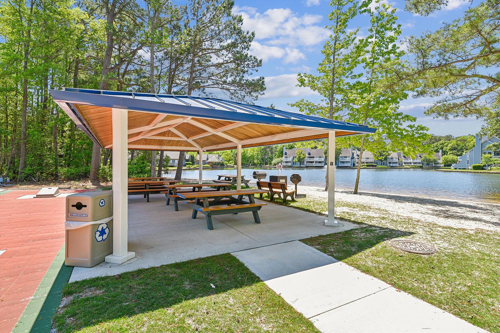 A covered pavilion with picnic tables sits beside a serene lake, surrounded by lush trees and a peaceful waterfront setting within the resort community.