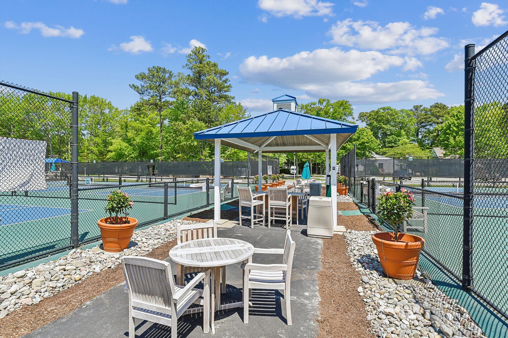 Outdoor recreation area features shaded seating pavilion between tennis courts with decorative planters and scenic tree backdrop under open blue skies.