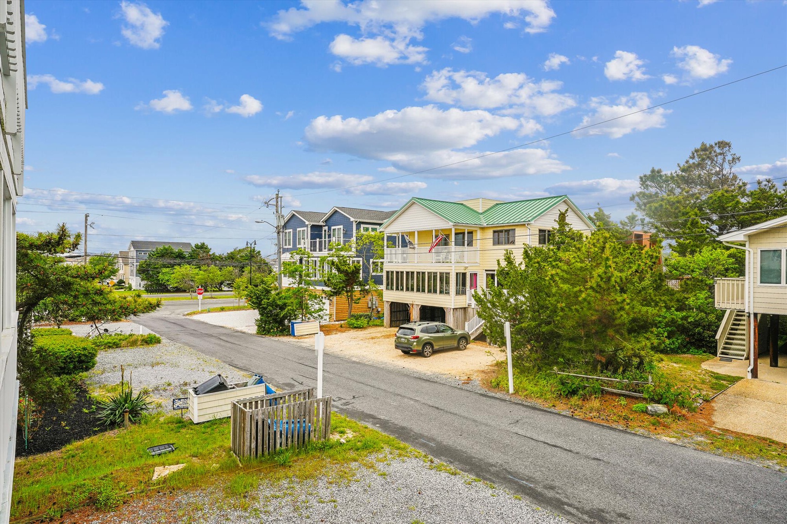 Bright yellow beach house with green metal roof nestled in a peaceful coastal neighborhood with mature trees and nearby vacation homes.
