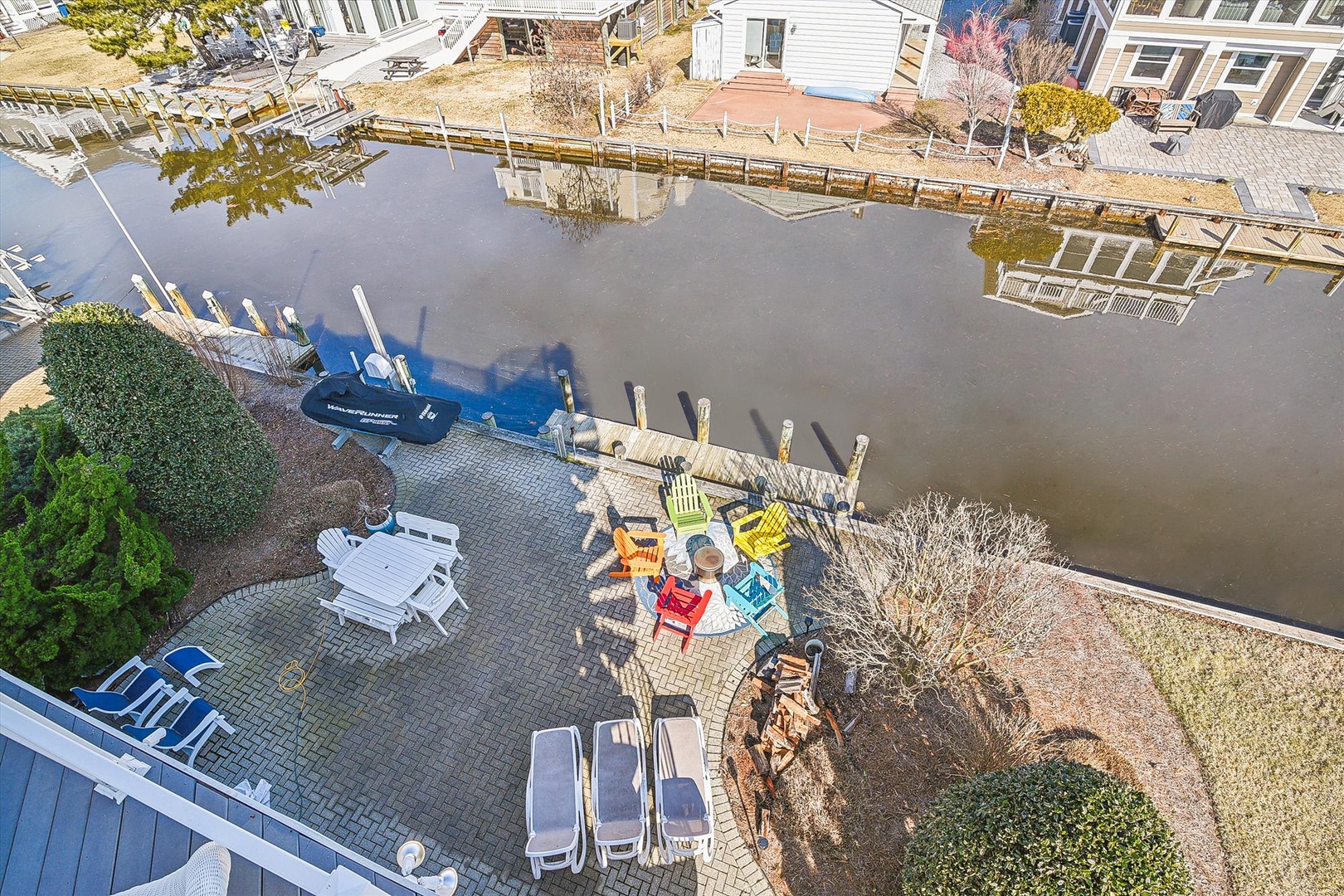 This aerial view captures the property's private waterfront location with direct canal access, a dock with boat slips, and a spacious patio featuring colorful Adirondack chairs around a firepit.