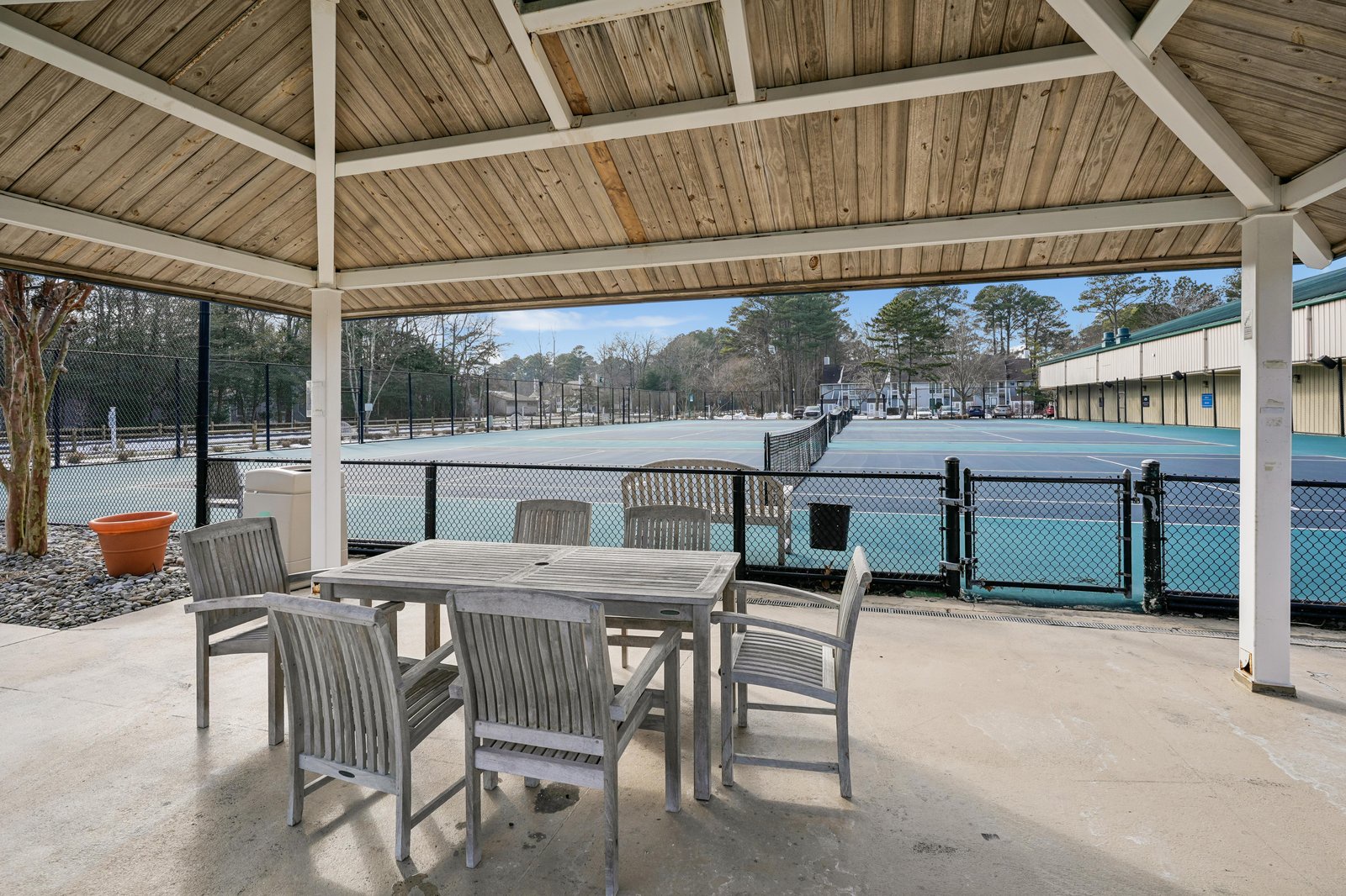 Covered patio seating area overlooks tennis courts with wooded backdrop. Seasonal outdoor gathering space near recreational facilities.
