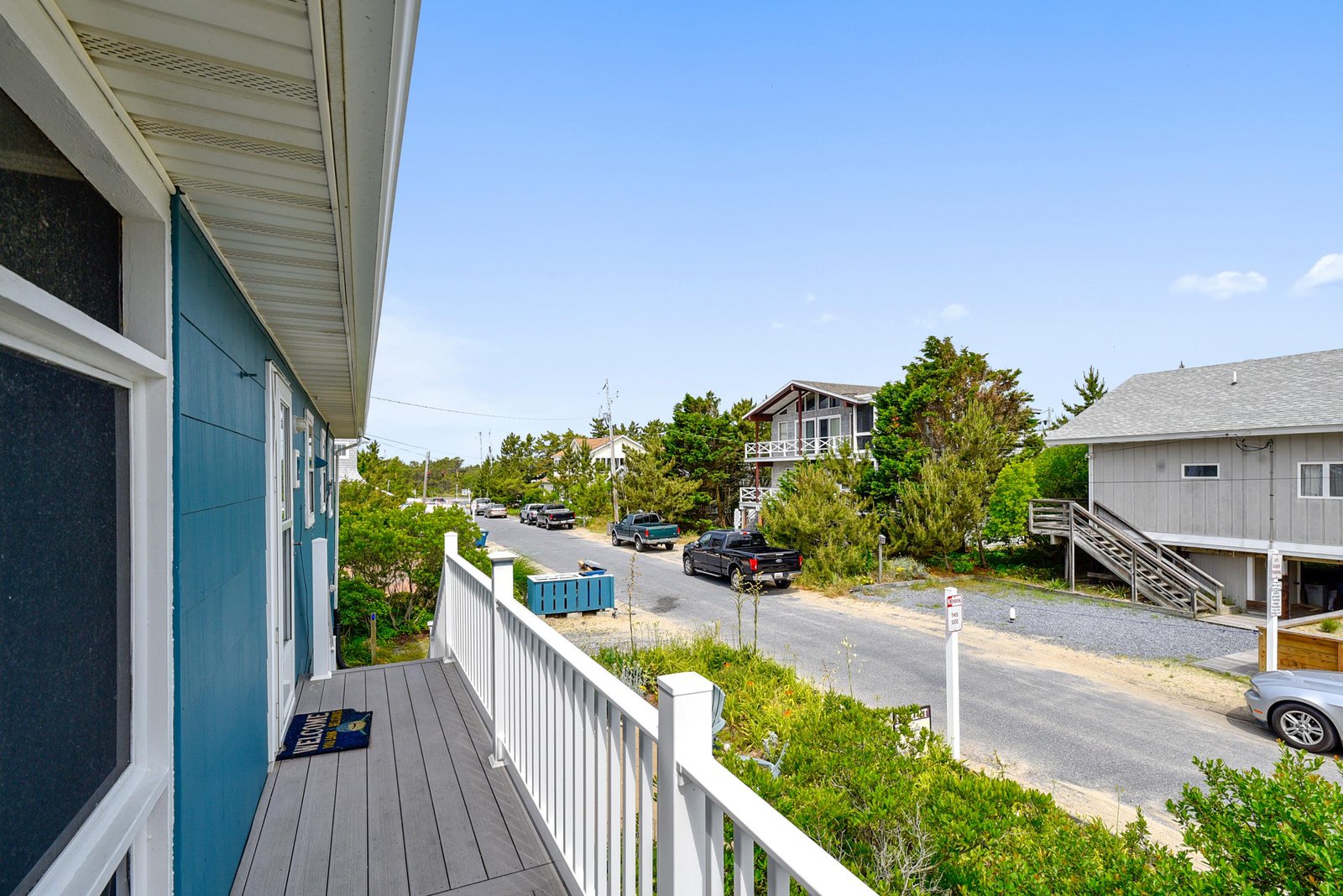 A bright summer day overlooks the quiet neighborhood street from the deck, with coastal homes and greenery framing the relaxed beach community atmosphere.