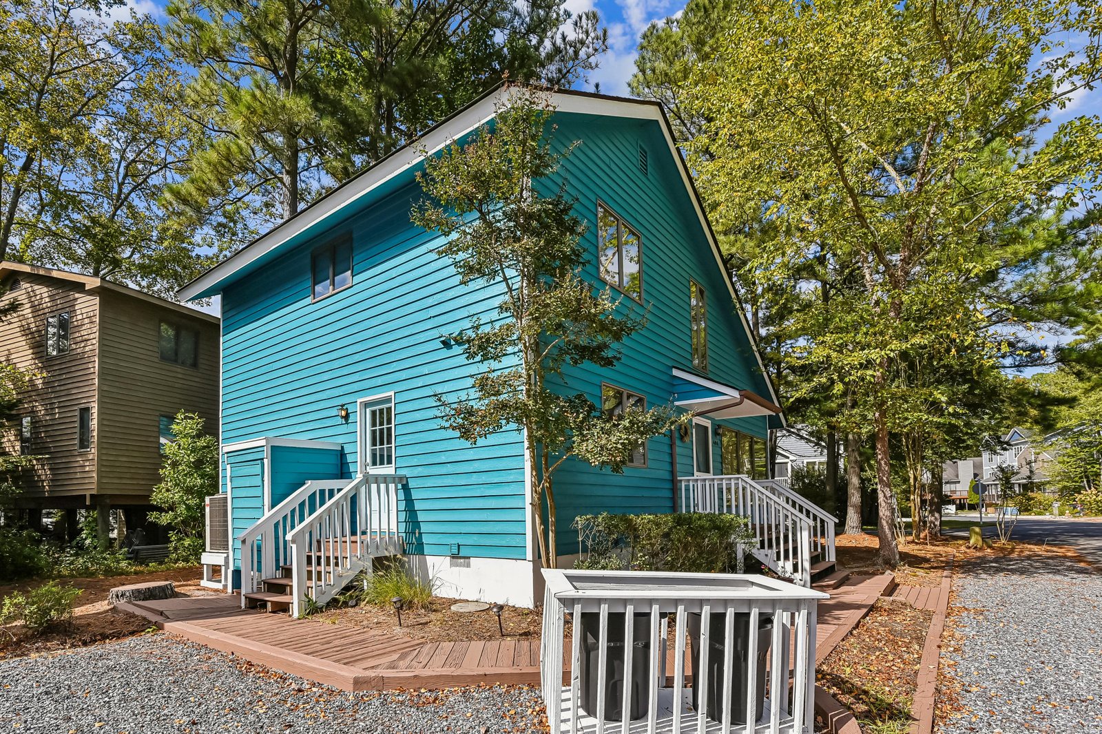 A charming turquoise cottage nestled among mature trees, offering a cheerful welcome with white railings and natural surroundings.
