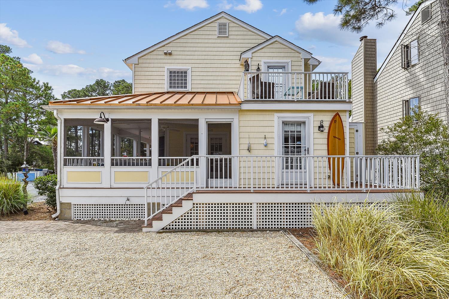 A charming coastal retreat with screened porch and upper deck, featuring inviting outdoor spaces for relaxation and gravel driveway parking, set among coastal greenery and neighboring beach homes.