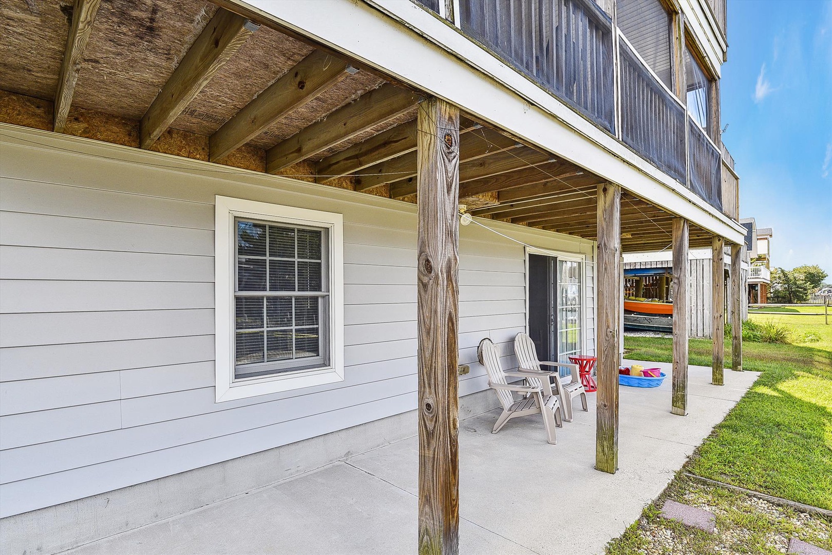 The covered patio with natural wood beams offers a shaded outdoor space perfect for relaxing in Adirondack chairs and enjoying the surrounding lawn.