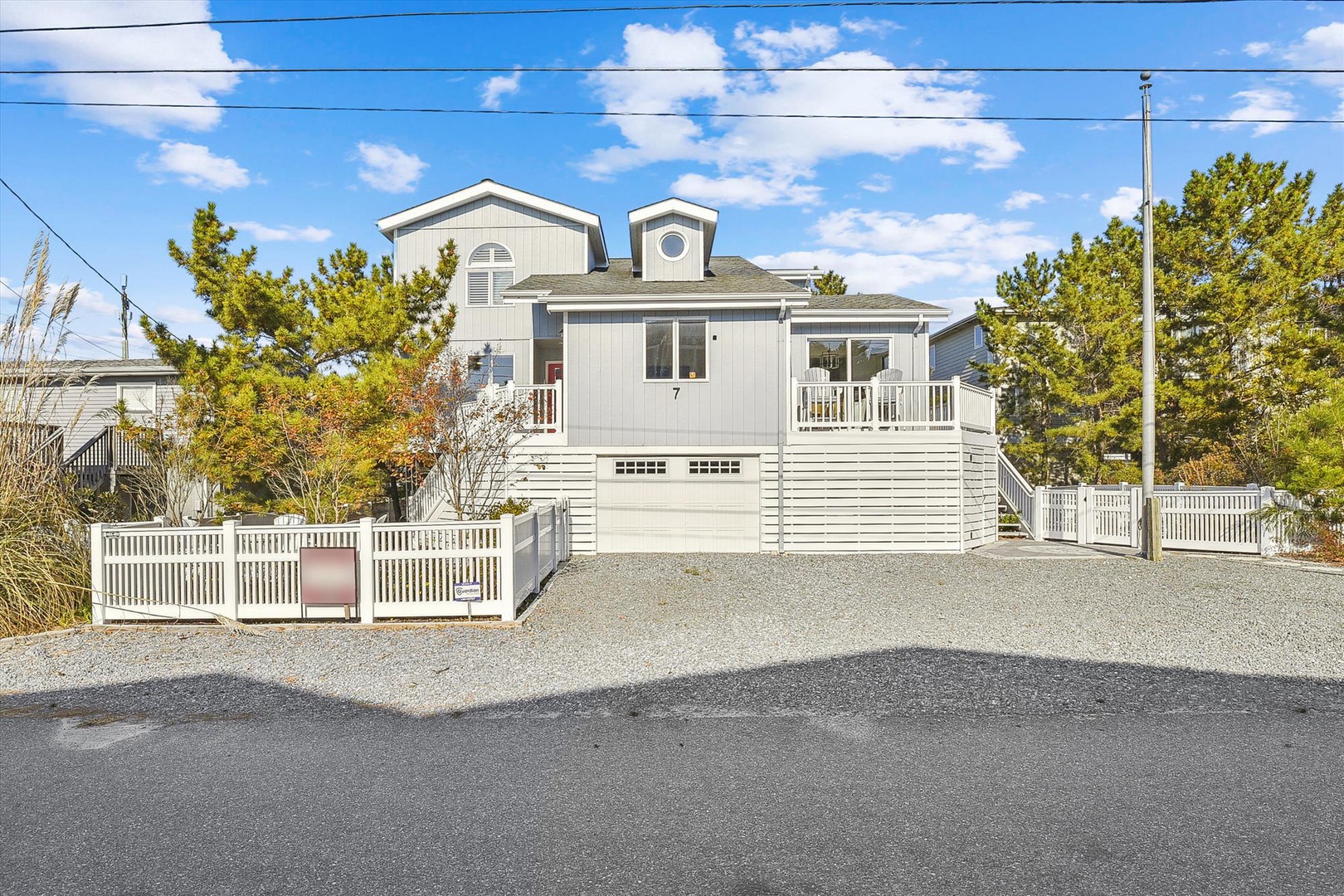 Coastal beach house with private parking and white picket fence, nestled among green trees in a quiet residential neighborhood near the shore.