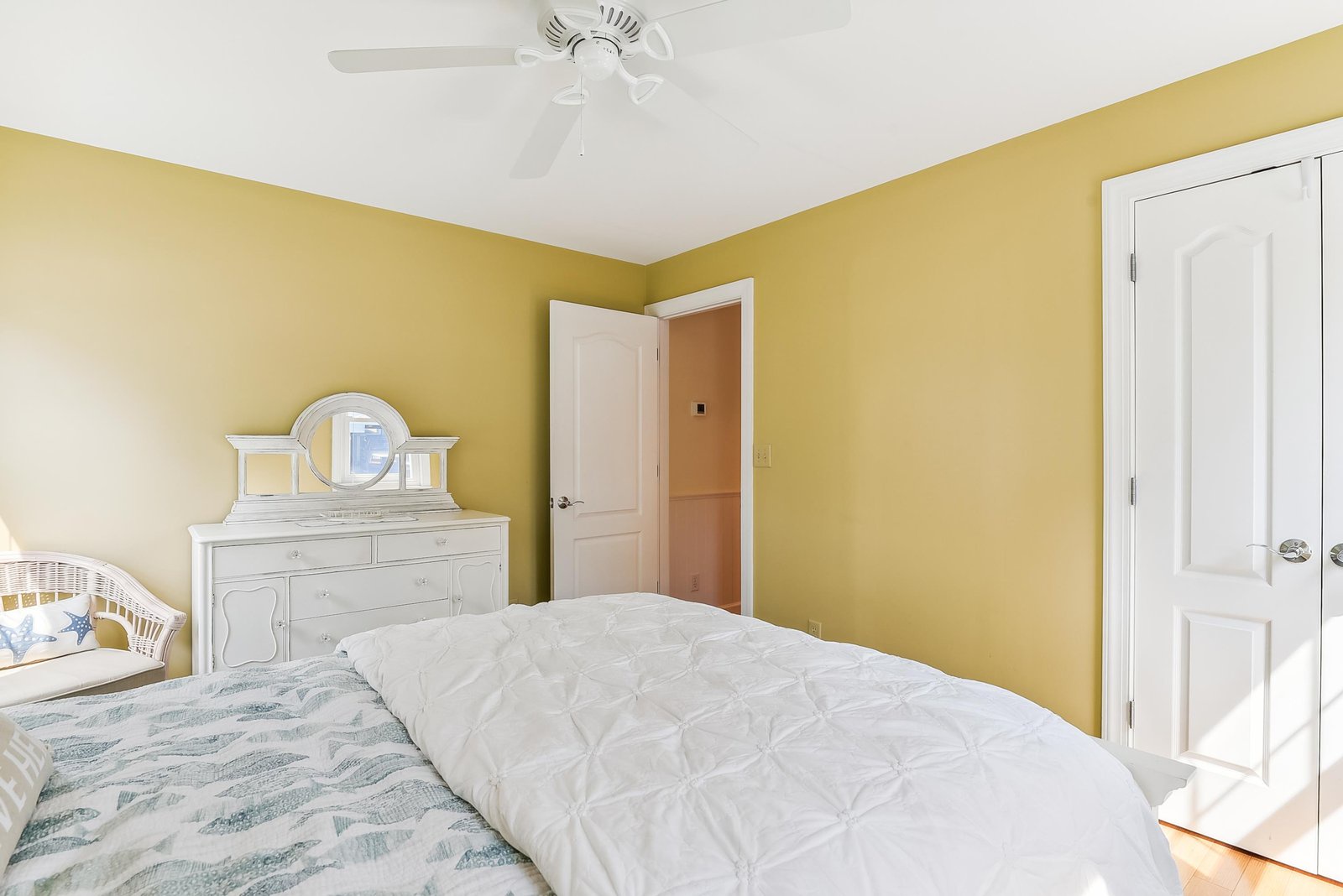 Step into your restful bedroom retreat with cheerful yellow walls, a ceiling fan keeping you comfortable, and a coastal-inspired bed inviting you to unwind.