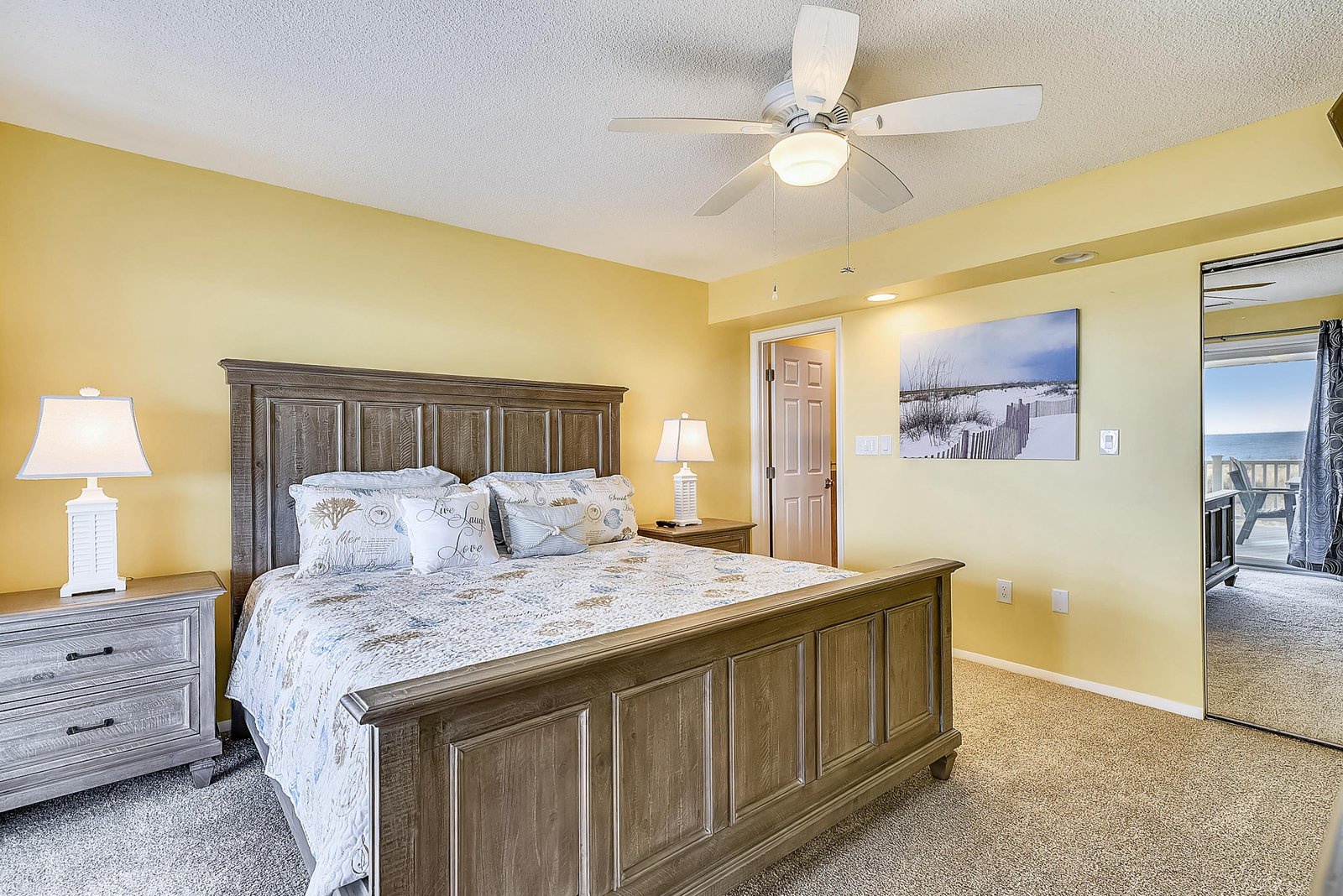 Your tranquil bedroom awaits with its soft yellow walls, plush bedding, and gentle ceiling fan. Drift off to the soothing coastal artwork and wake to ocean breezes from your private balcony.