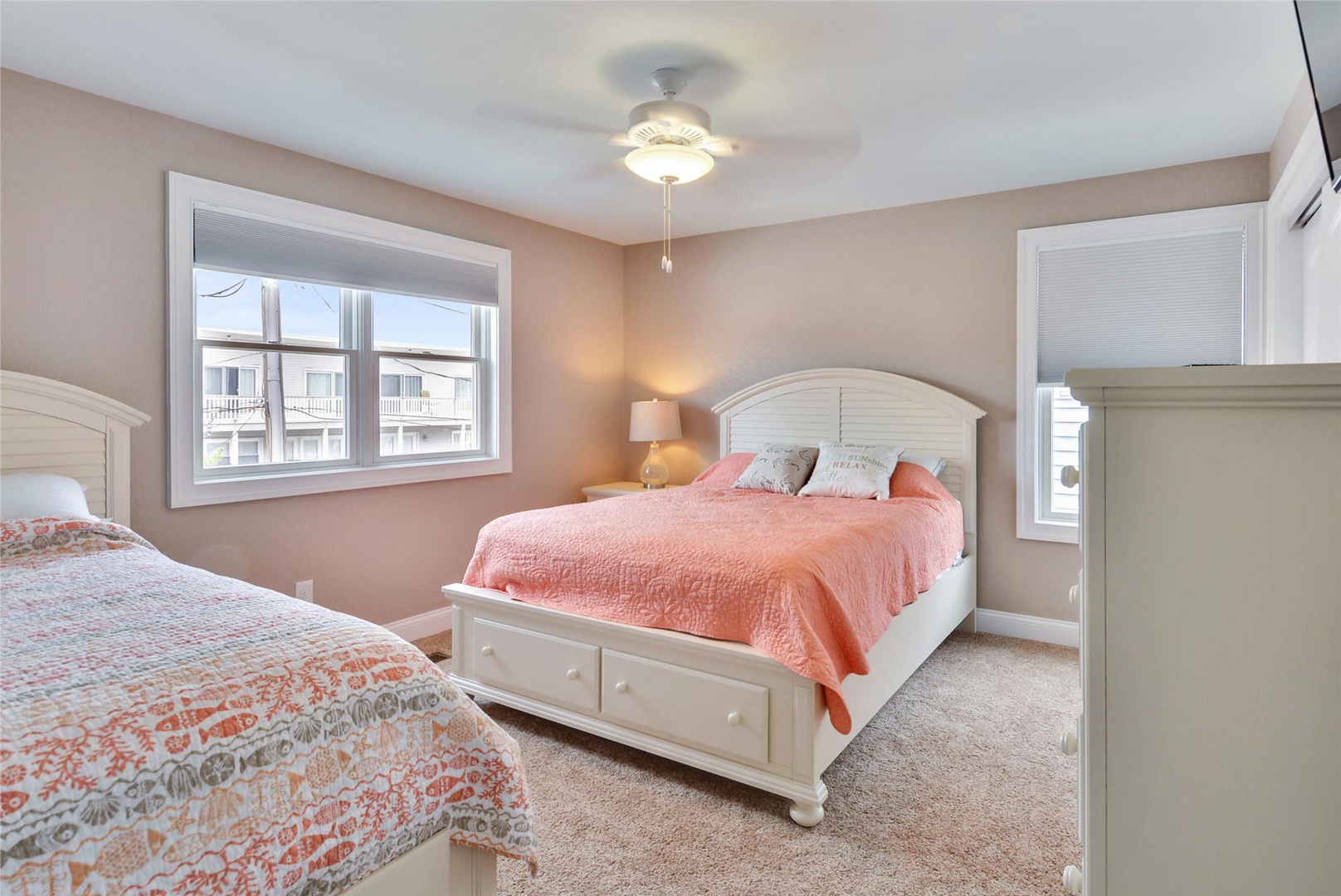 Your coastal-style bedroom awaits with two plush beds dressed in soft peach tones, gentle ceiling fan breeze, and warm natural light streaming through the windows for your peaceful retreat.