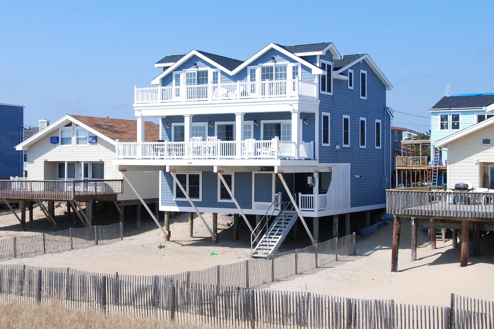 Beachfront property with blue coastal siding, white trim, multiple balconies, and direct beach access via a private stairway leading to sandy shores. Neighboring properties visible.