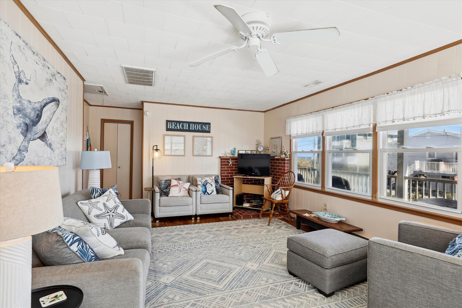 Your coastal retreat awaits with a spacious living room featuring comfortable seating, beachy décor, and large windows framing neighborhood views. Relax under the ceiling fan in this inviting space perfect for gathering after beach days.