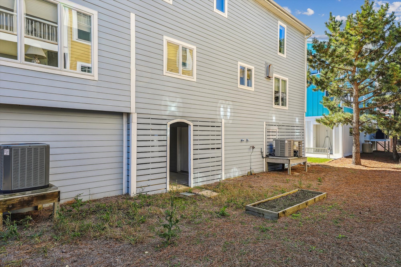 The property's exterior showcases a quiet residential setting with air conditioning units, ample natural light, and a spacious, private backyard area bordered by pine trees.