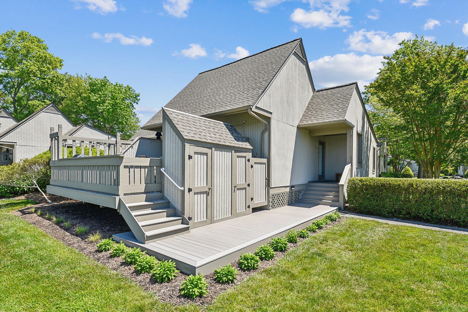 A charming modern farmhouse-style rental with vertical siding, nestled in a peaceful residential community with mature trees, manicured lawns, and welcoming outdoor deck space.