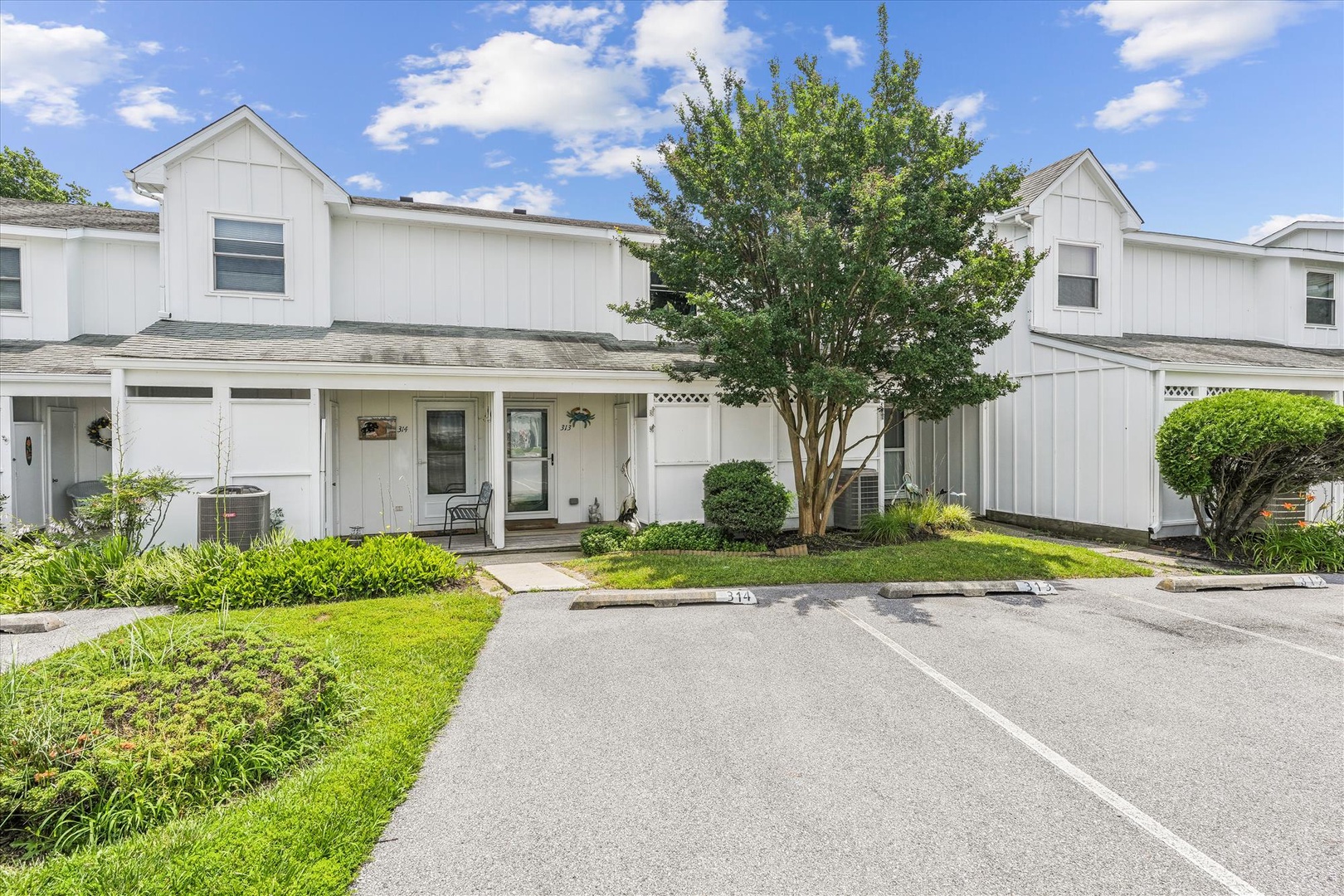 A modern two-story townhome with white siding, mature landscaping, and dedicated parking in a peaceful residential setting with blue skies overhead.
