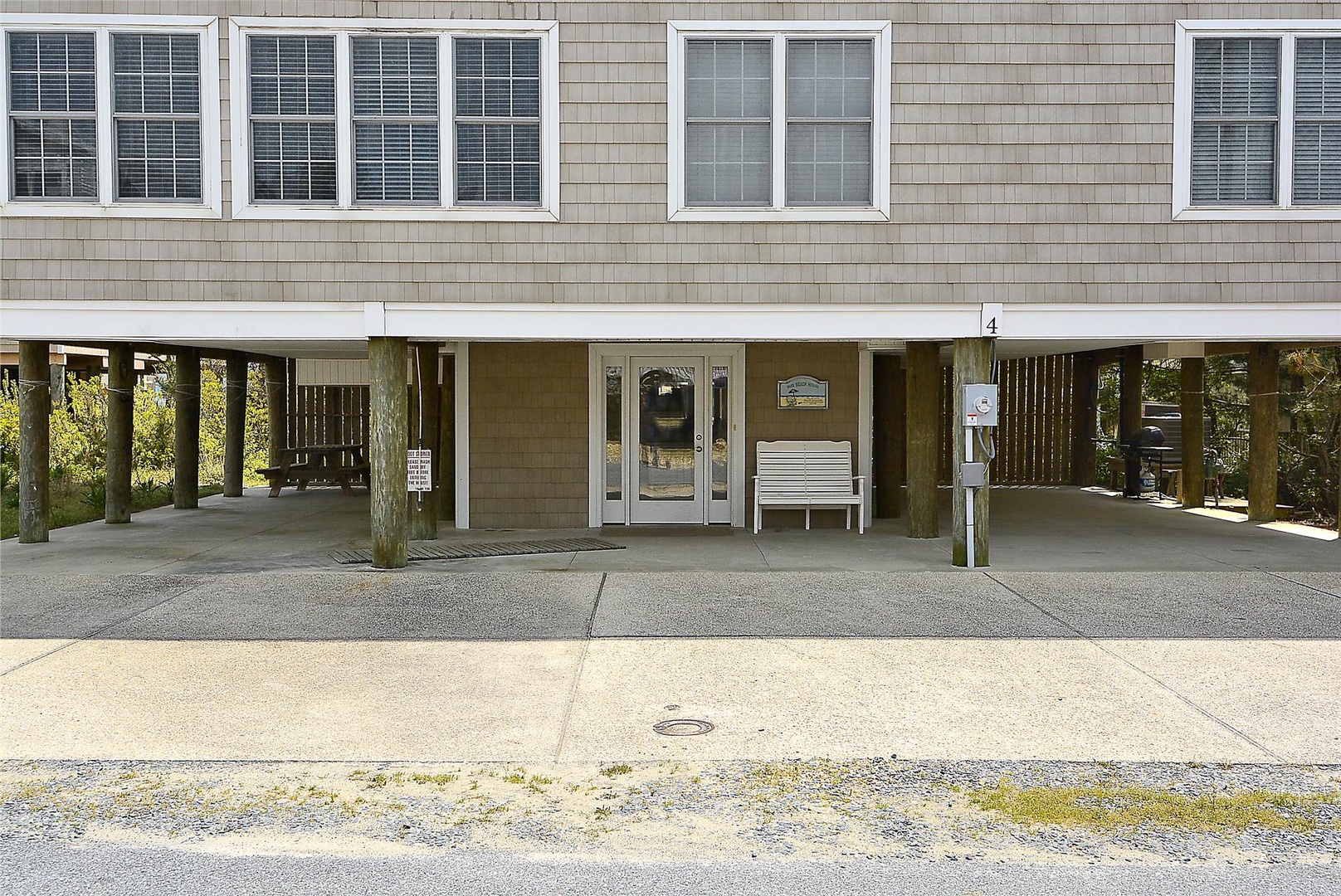 Exterior view of a raised beach house featuring covered ground-level parking, a prominent front entrance with glass doors, and neutral shingle siding. The property is elevated on pilings common to coastal architecture.