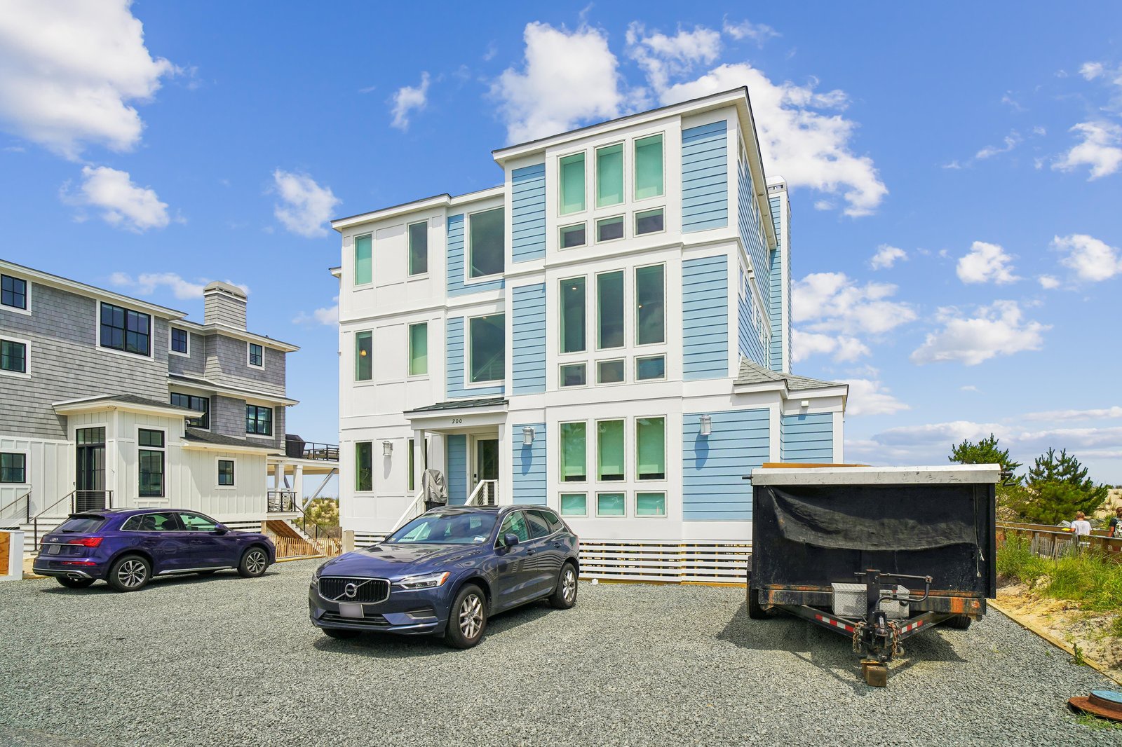 A modern three-story coastal property with bright blue and white siding, featuring private parking and neighboring residences in a peaceful beachside setting.