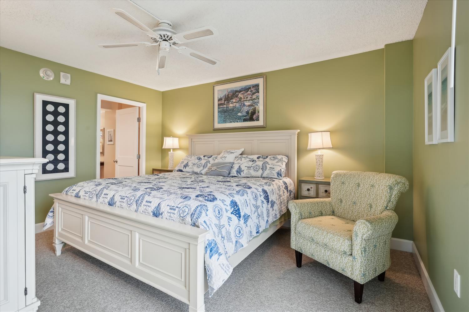 Your serene bedroom retreat features a plush king bed with coastal linens, a cozy reading chair, and calming olive-green walls that invite restful nights and peaceful mornings.