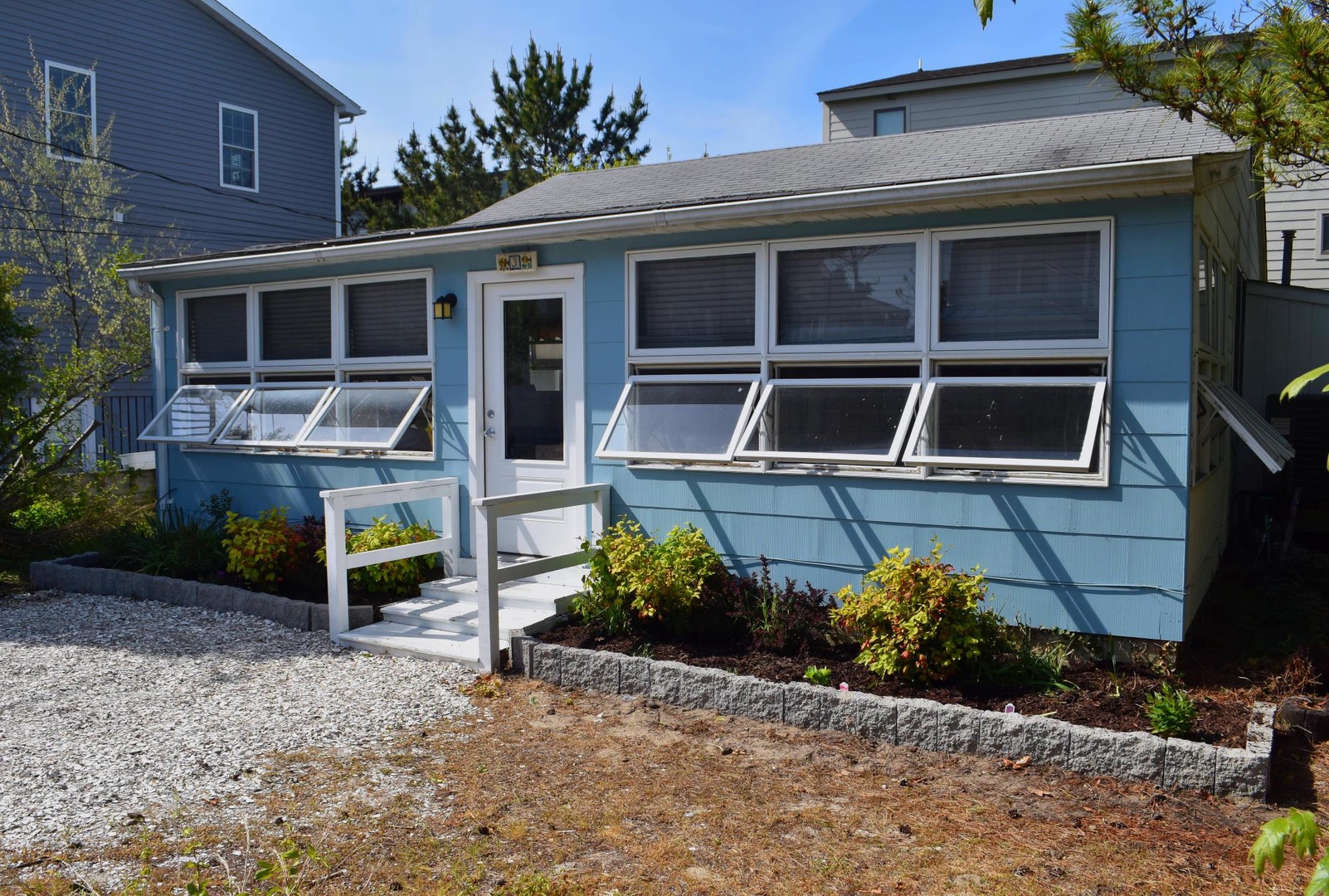A charming blue cottage with white trim and cheerful flower beds welcomes you to your seaside getaway. The property features a gravel parking area, cozy front porch, and is nestled in a quiet residential neighborhood.
