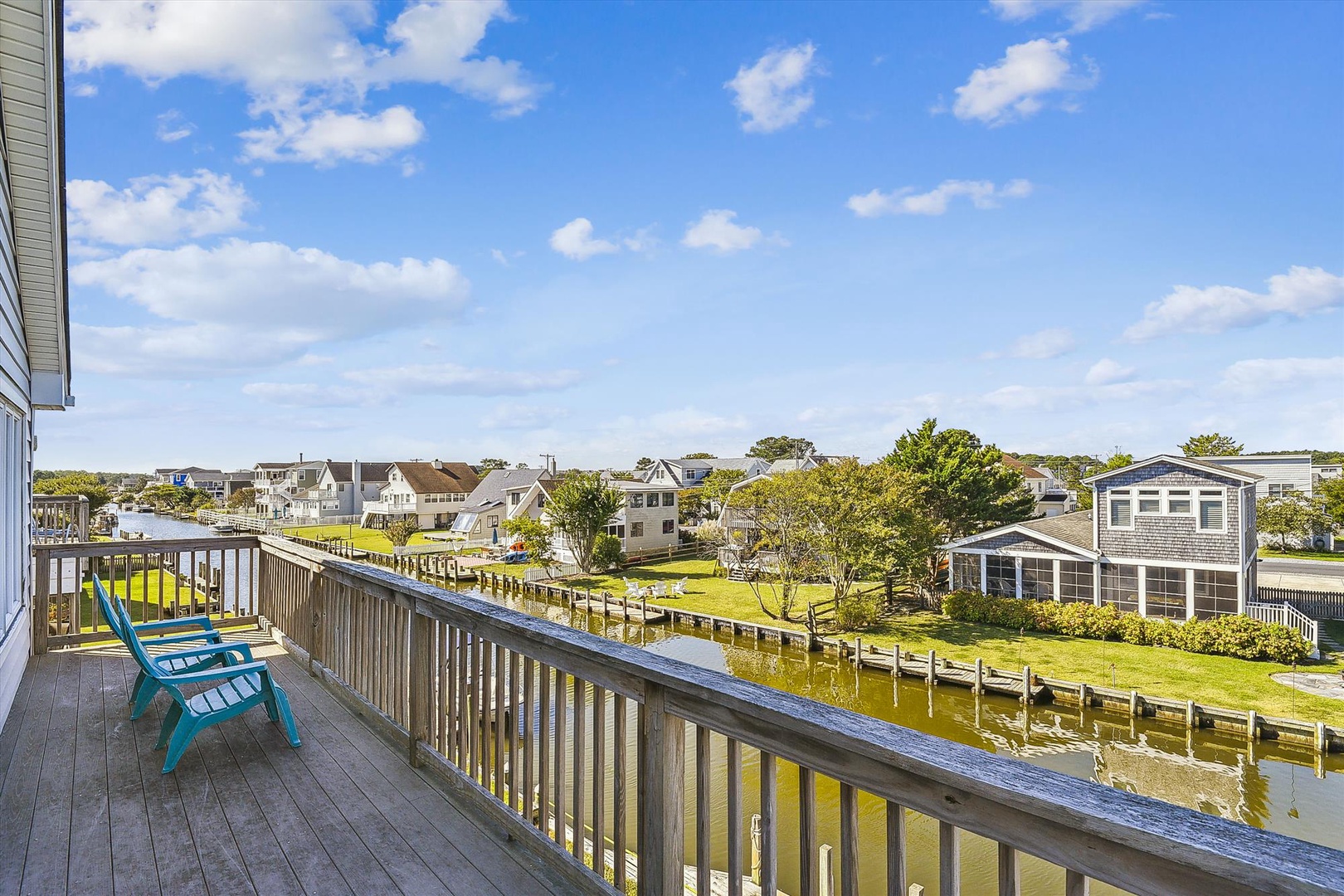 A peaceful waterfront neighborhood unfolds beyond this balcony, where homes and docks line the calm canal under a bright blue sky dotted with clouds.