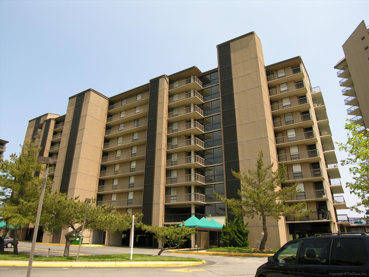 Multi-story building features private balconies for every unit, situated in a landscaped area with convenient street parking and green awnings marking the main entrance.