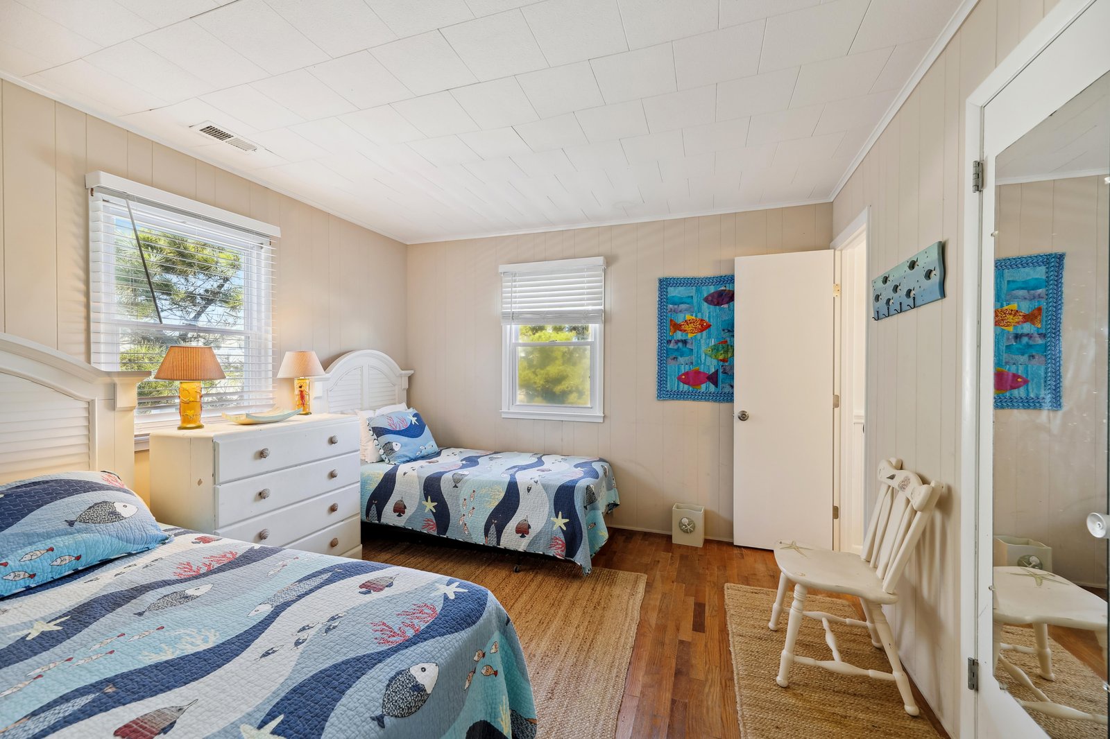 Bright and cheerful bedroom with twin beds dressed in ocean-themed quilts, warm wood floors, and sunlight streaming through the windows. Your kids will love the playful fish decor and cozy coastal charm.