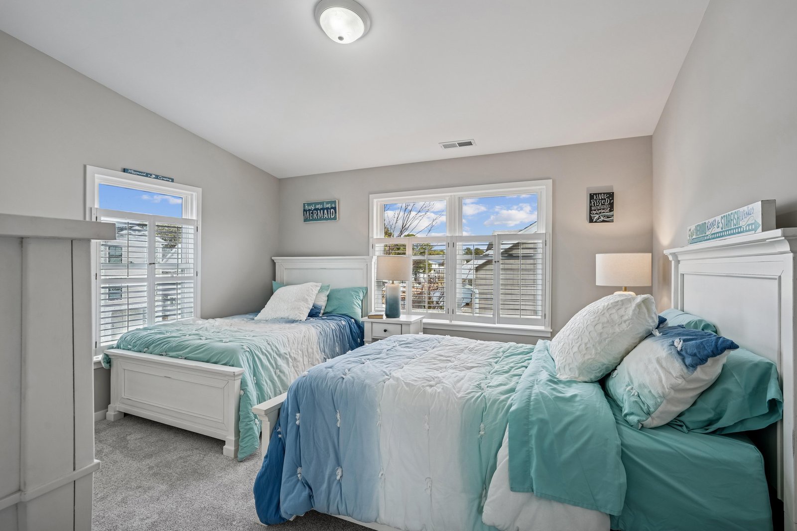 Your bright coastal bedroom with two cozy beds dressed in seafoam and aqua tones. Wake to sunshine streaming through plantation shutters and enjoy peaceful neighborhood views from your spacious windows.