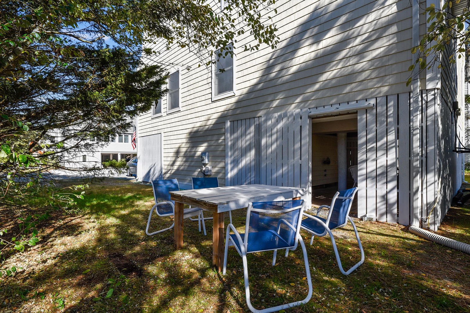 The outdoor dining area sits in a shaded garden space with a simple wooden table and comfortable chairs, offering a quiet spot near the property entrance for meals or relaxation.