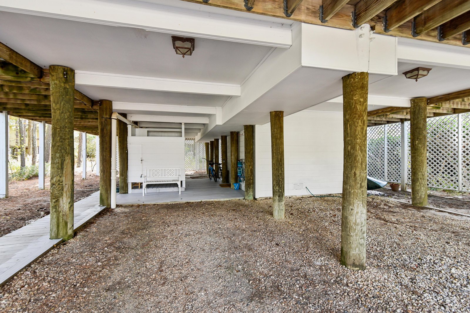 Elevated coastal property with spacious covered parking and storage area on gravel foundation, surrounded by natural wooded setting and lattice privacy screens.