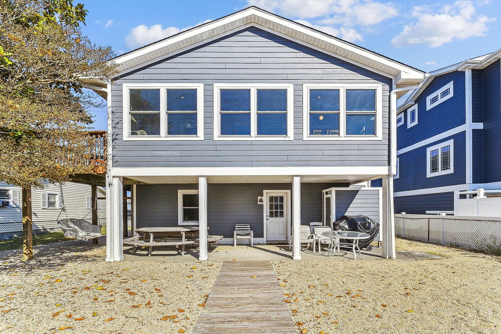 Charming beach house exterior featuring shaded outdoor seating and dining areas beneath the elevated upper floor, with a grill ready for coastal cookouts and neighboring properties visible in the surrounding residential area.
