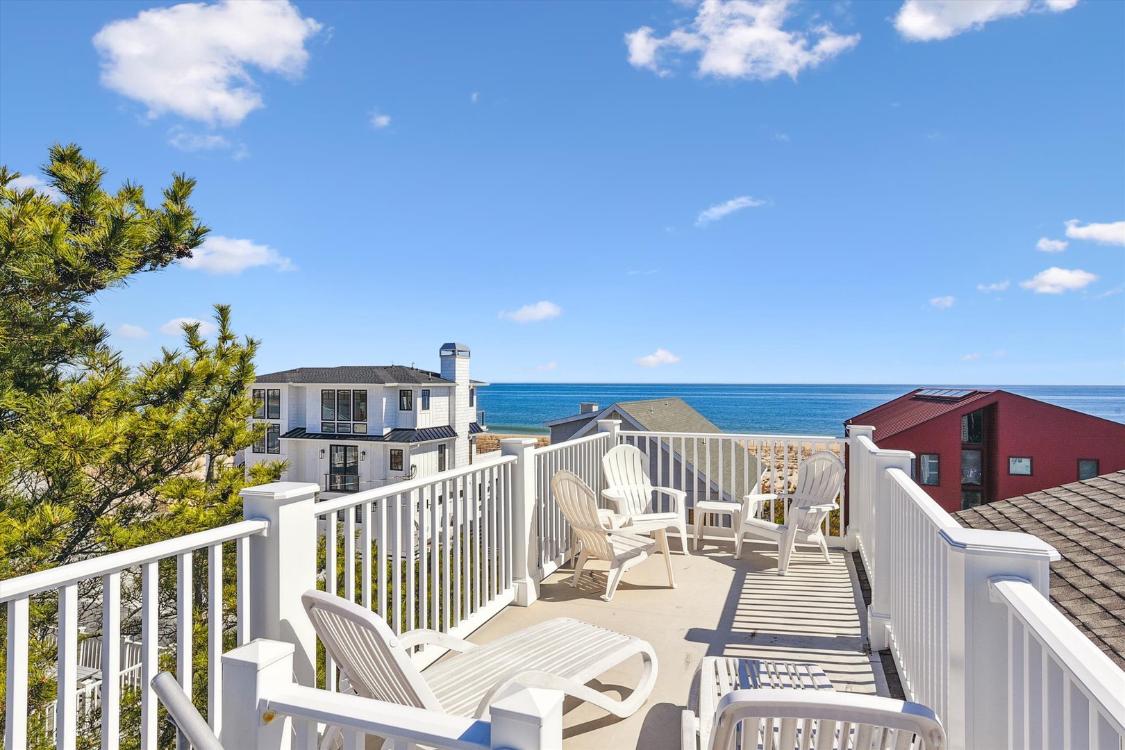 A sun-drenched rooftop deck features ample seating and sweeping coastal views overlooking the ocean, neighboring beach homes, and clear blue skies.
