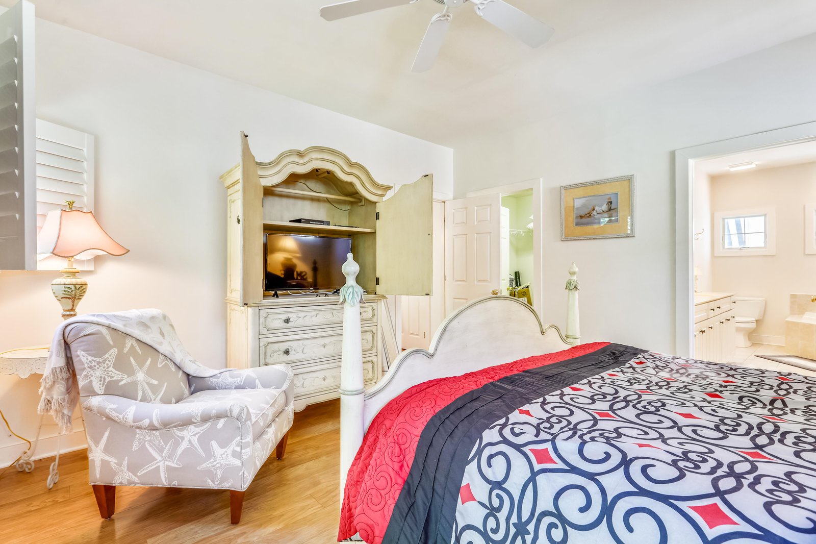 Your spacious bedroom invites you to unwind with a plush bed dressed in vibrant linens, a cozy armchair for quiet moments, and an elegant armoire revealing your entertainment center. Warm wood floors and soft coastal touches create a serene retreat.