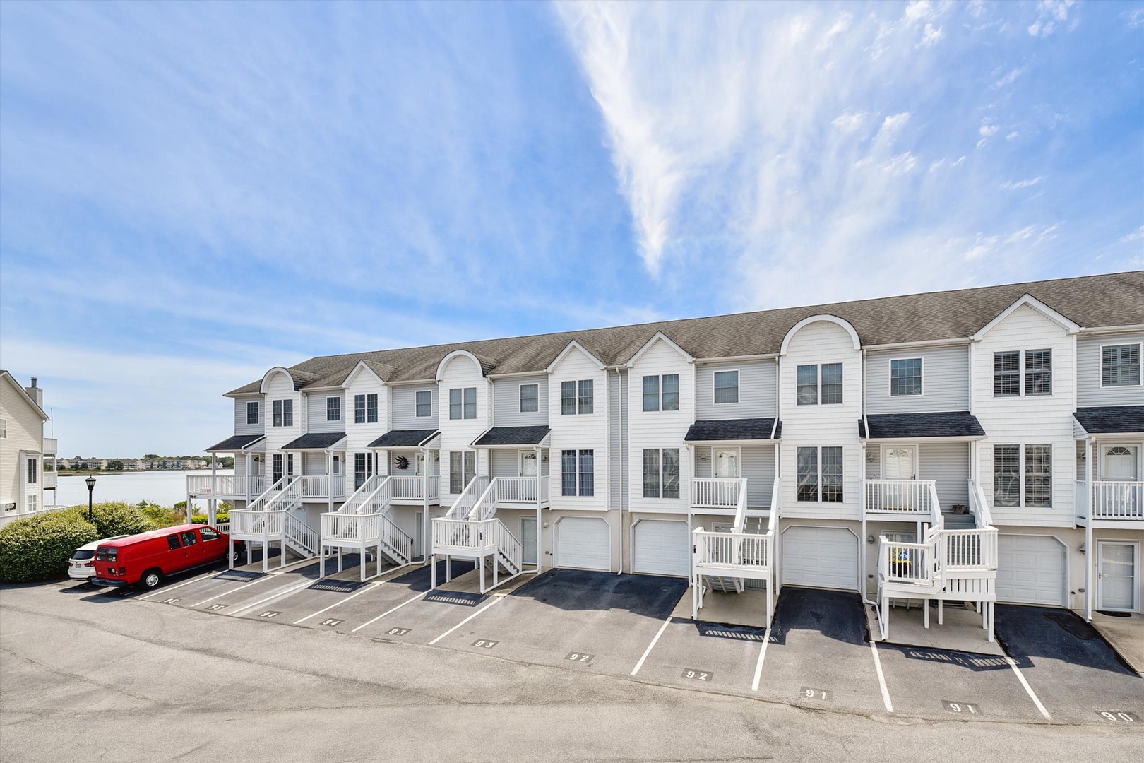A row of white townhomes with elevated entrances and individual garages, surrounded by parking spaces and landscaped greenery under a bright blue sky.