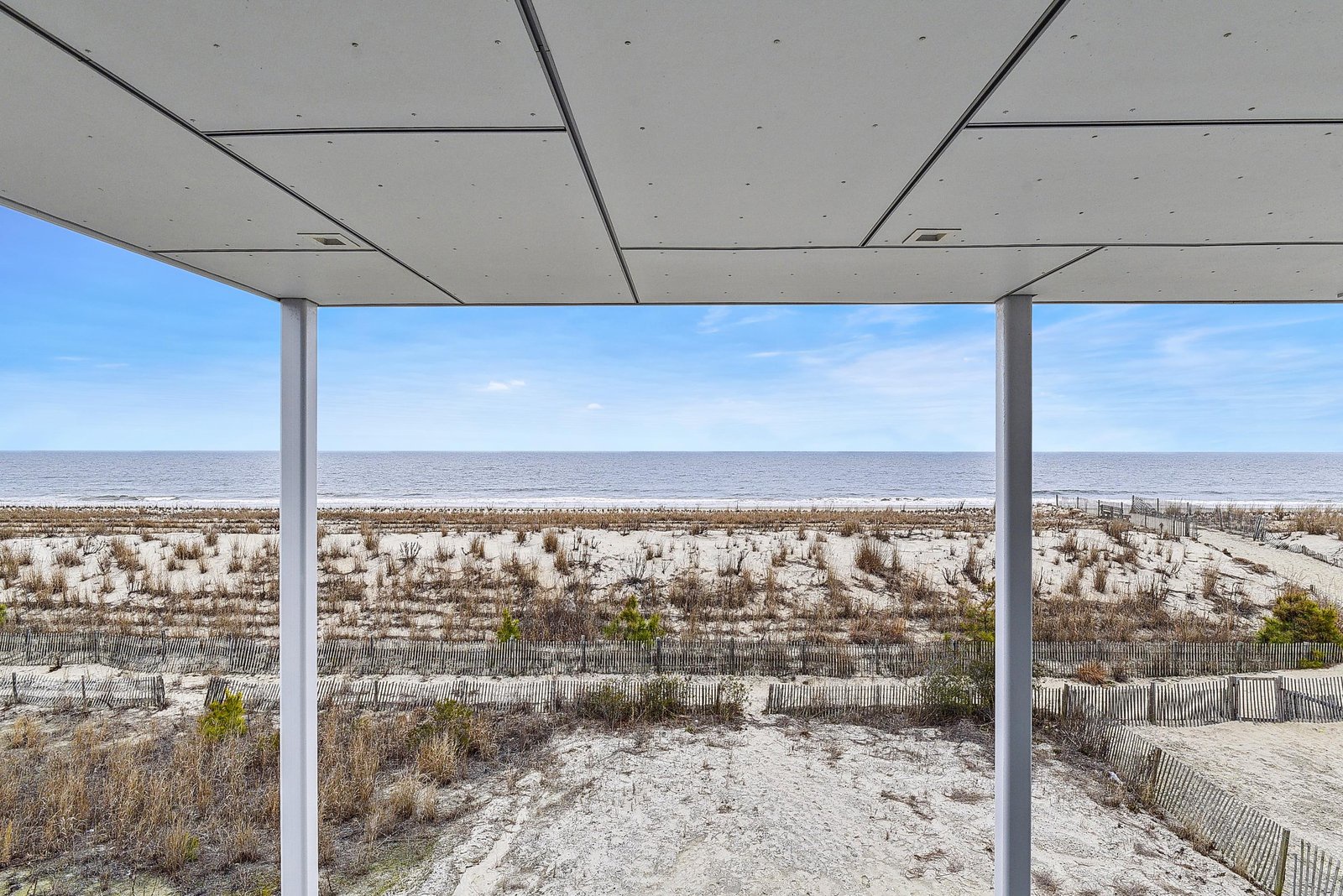 A covered terrace frames uninterrupted views of sandy dunes and the sparkling ocean beyond, where gentle waves meet endless sky.