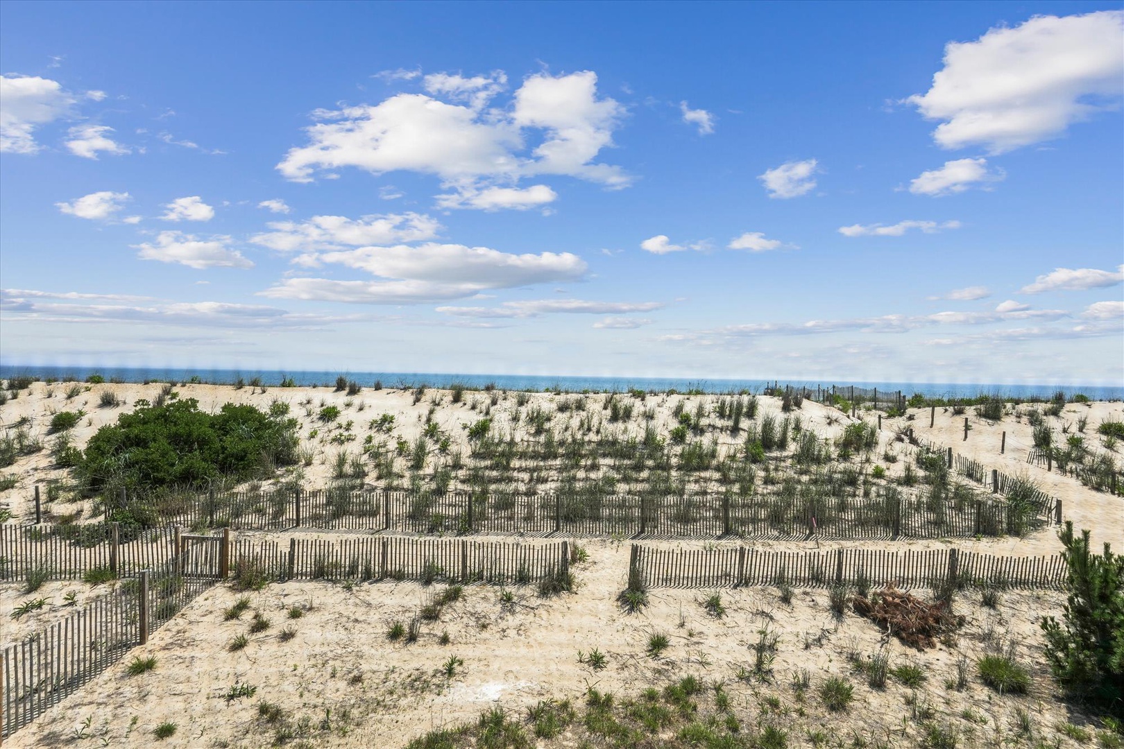Scenic coastal dunes bordered by weathered fencing lead toward sparkling blue waters under a bright, cloud-streaked sky—part of the property's surrounding natural landscape.