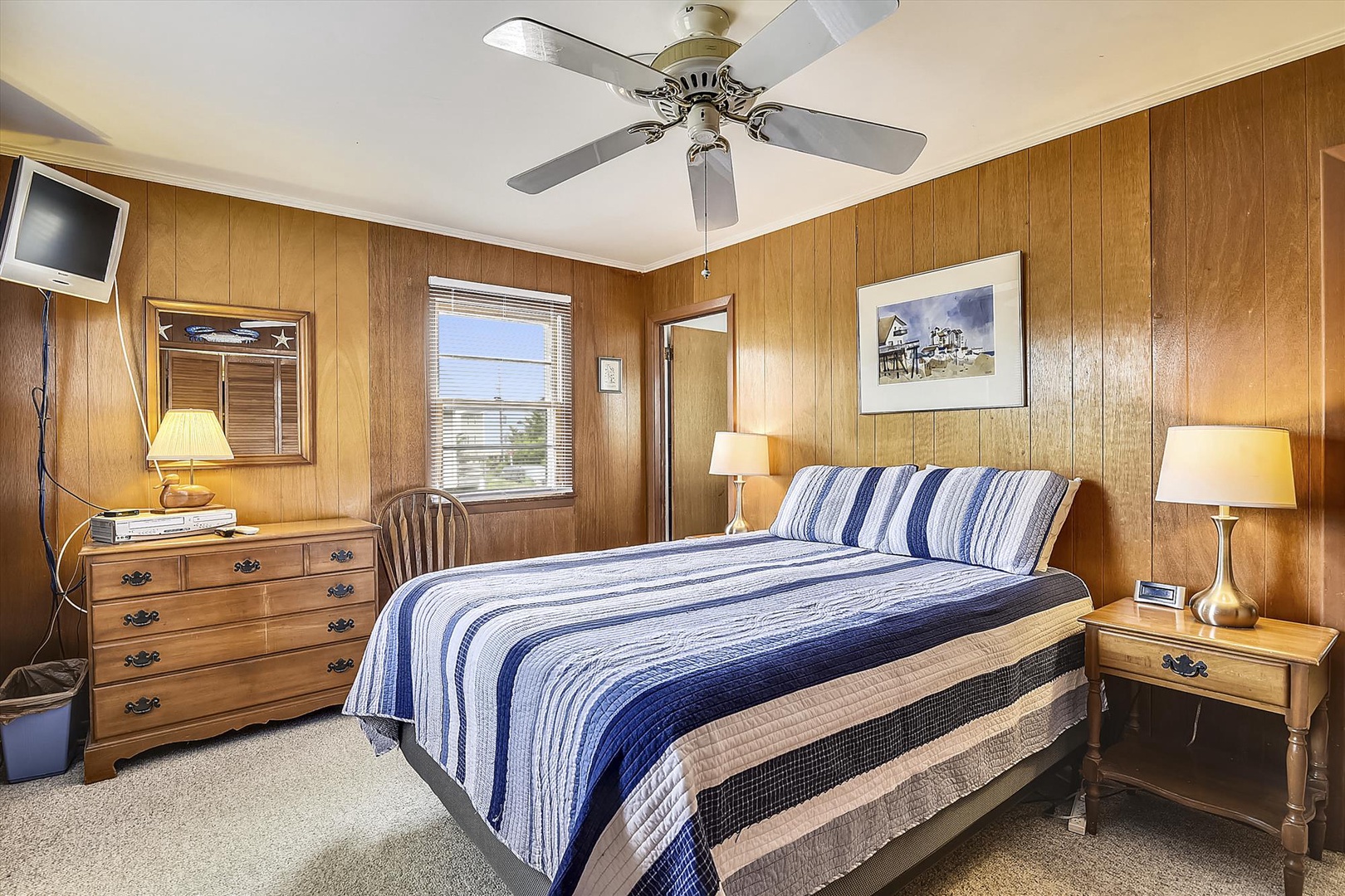 Unwind in this cozy bedroom where warm wood paneling and nautical-striped bedding create your peaceful coastal retreat, complete with a ceiling fan for comfort.