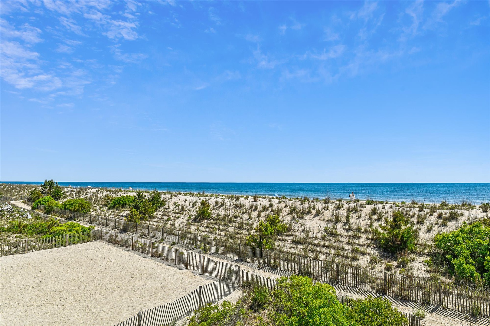 Sandy pathway leads through coastal dunes to pristine beach and turquoise waters under clear blue skies. Nearby natural landscape offers peaceful seaside setting.
